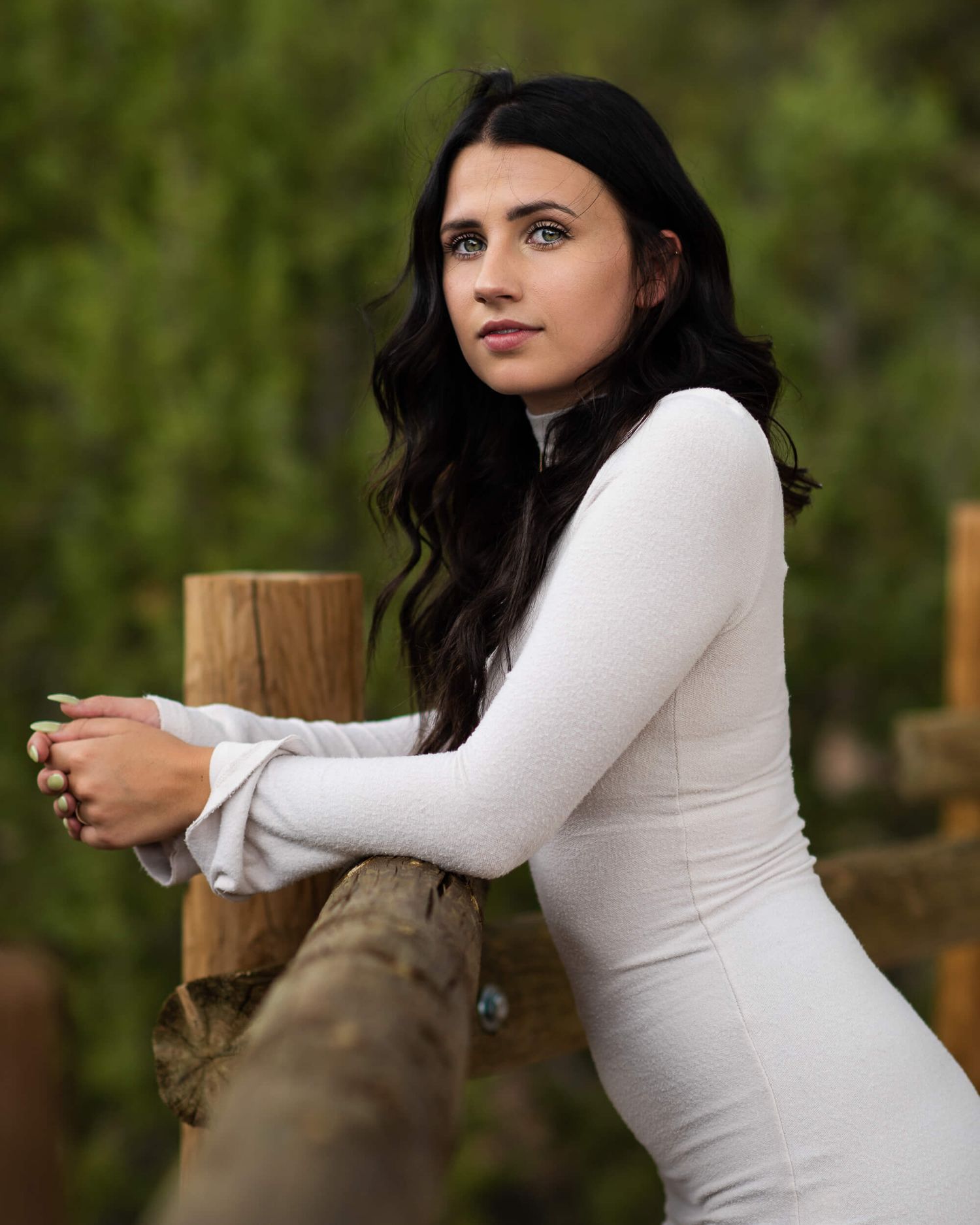 Young woman in white long-sleeve dress leaning on rustic wooden fence with green trees in background.