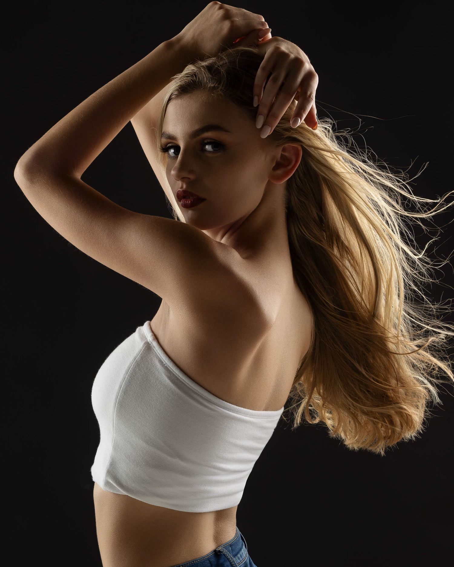 Young blonde woman in white crop top and jeans poses against dark background with hair flowing.