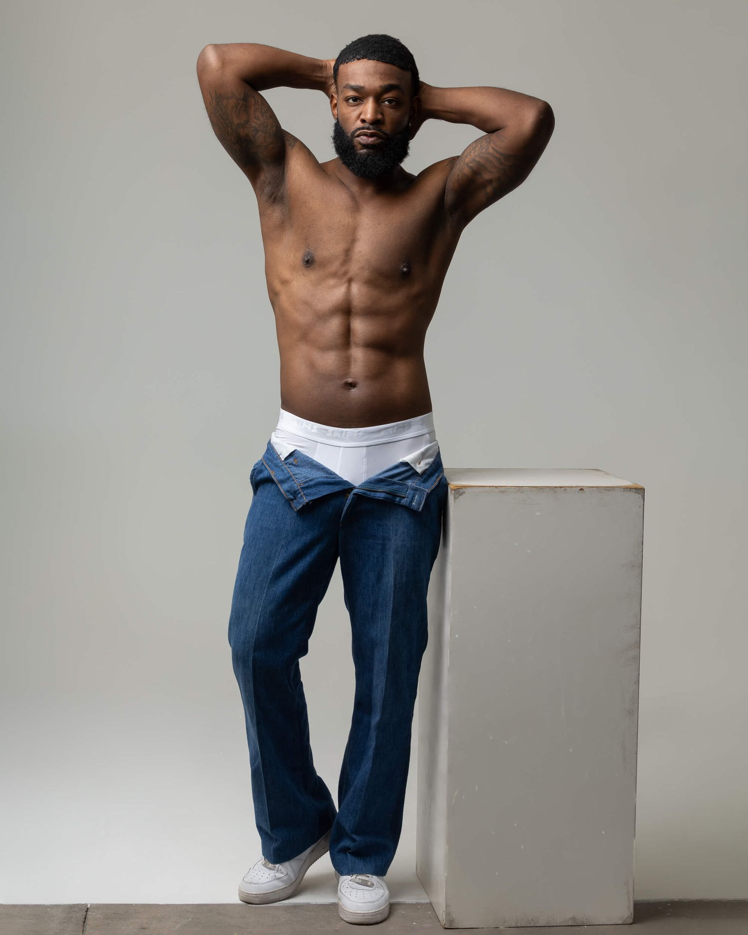 Shirtless muscular man with beard poses confidently in blue jeans and white sneakers against neutral background.