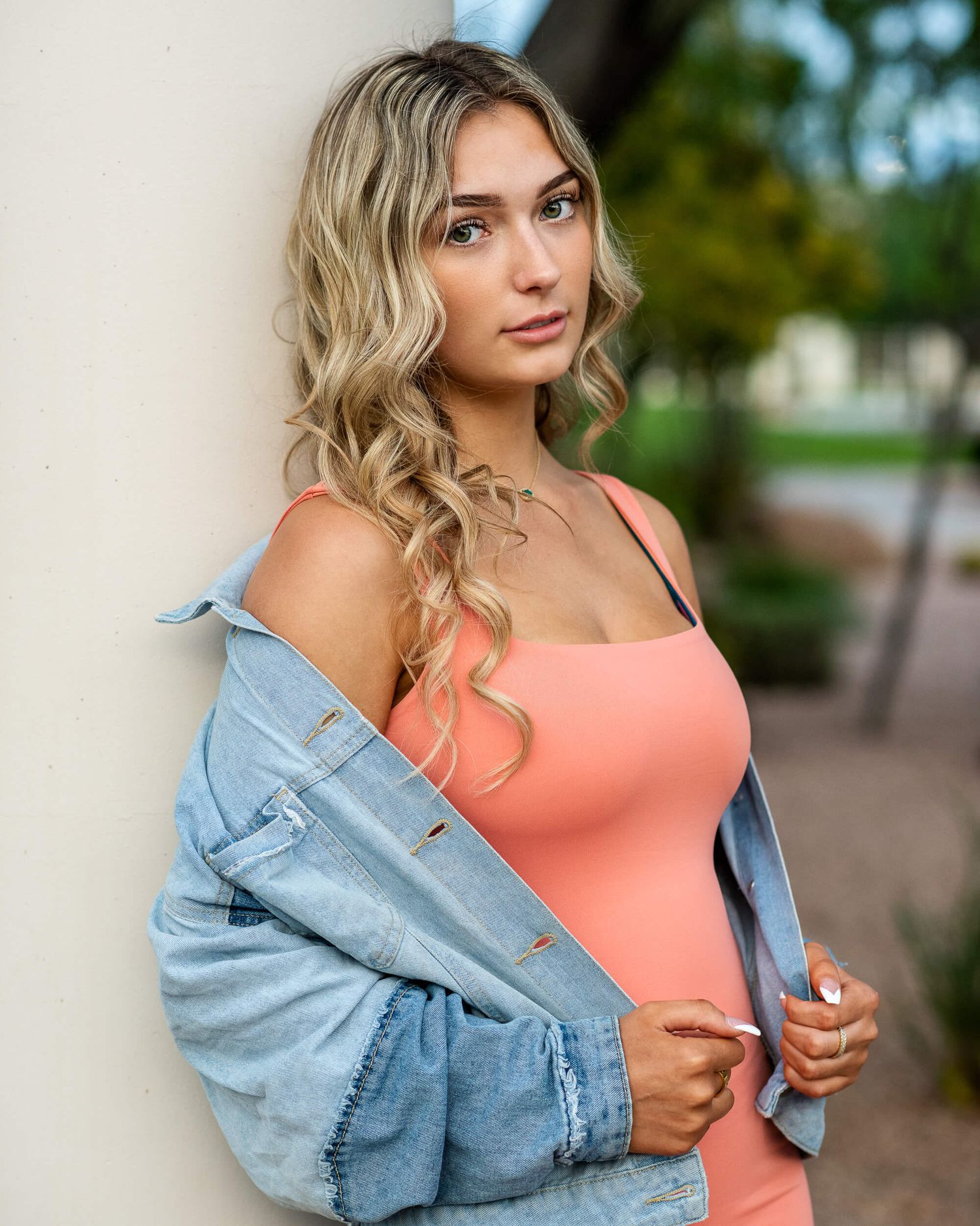 Blonde woman in peach bodysuit and denim jacket leaning against white pillar outdoors.