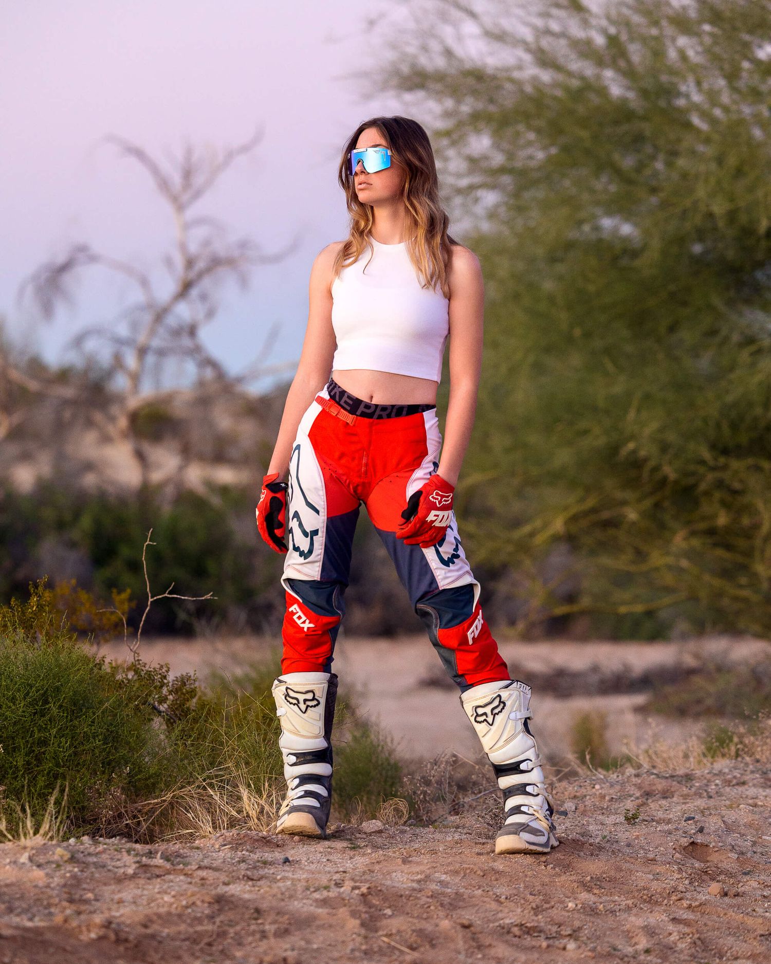 Woman in Fox Racing motocross pants and white crop top standing on desert trail.