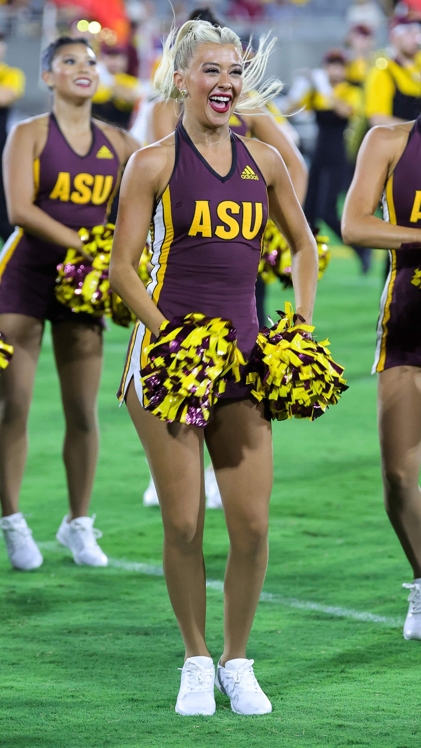 ASU cheerleader in maroon uniform holds gold pom-poms while performing on football field.