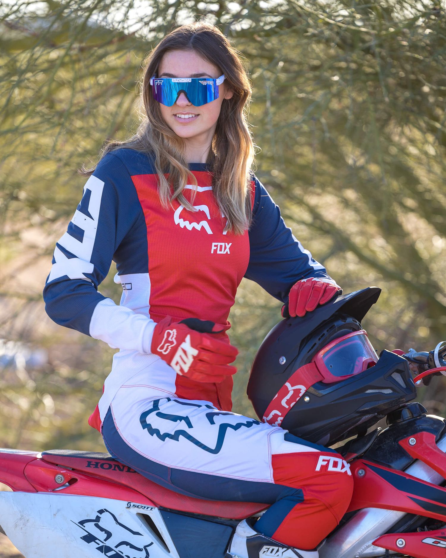 Female motocross rider in Fox Racing gear posing with a Honda dirt bike outdoors.