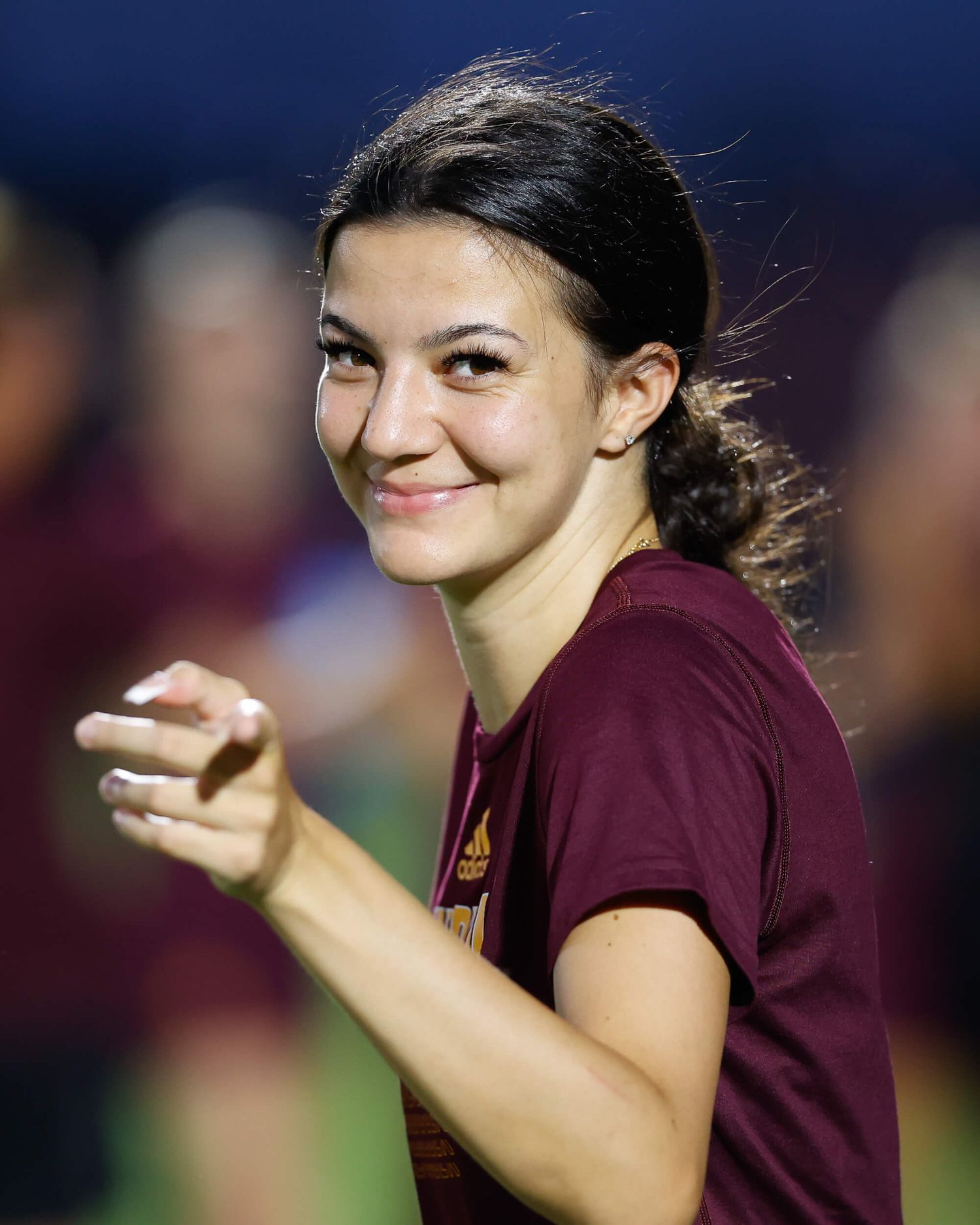Smiling young woman in maroon athletic shirt gestures at outdoor sports event.