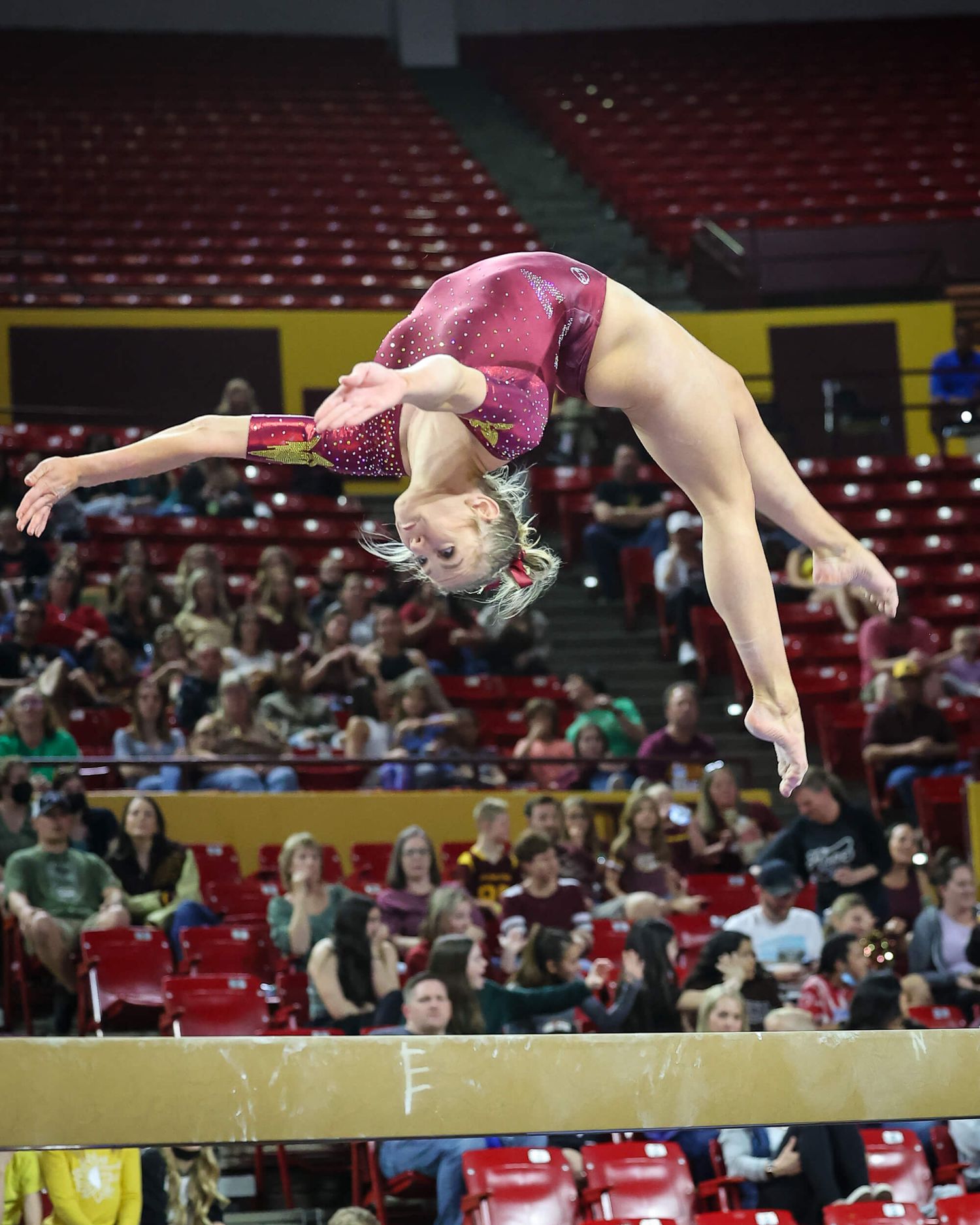 Gymnast performs back walkover on balance beam before stadium crowd.