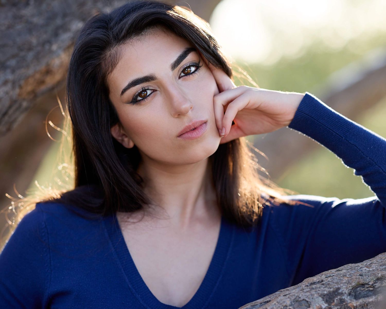 Brunette woman in a blue v-neck sweater posing outdoors in golden hour light for a portrait.