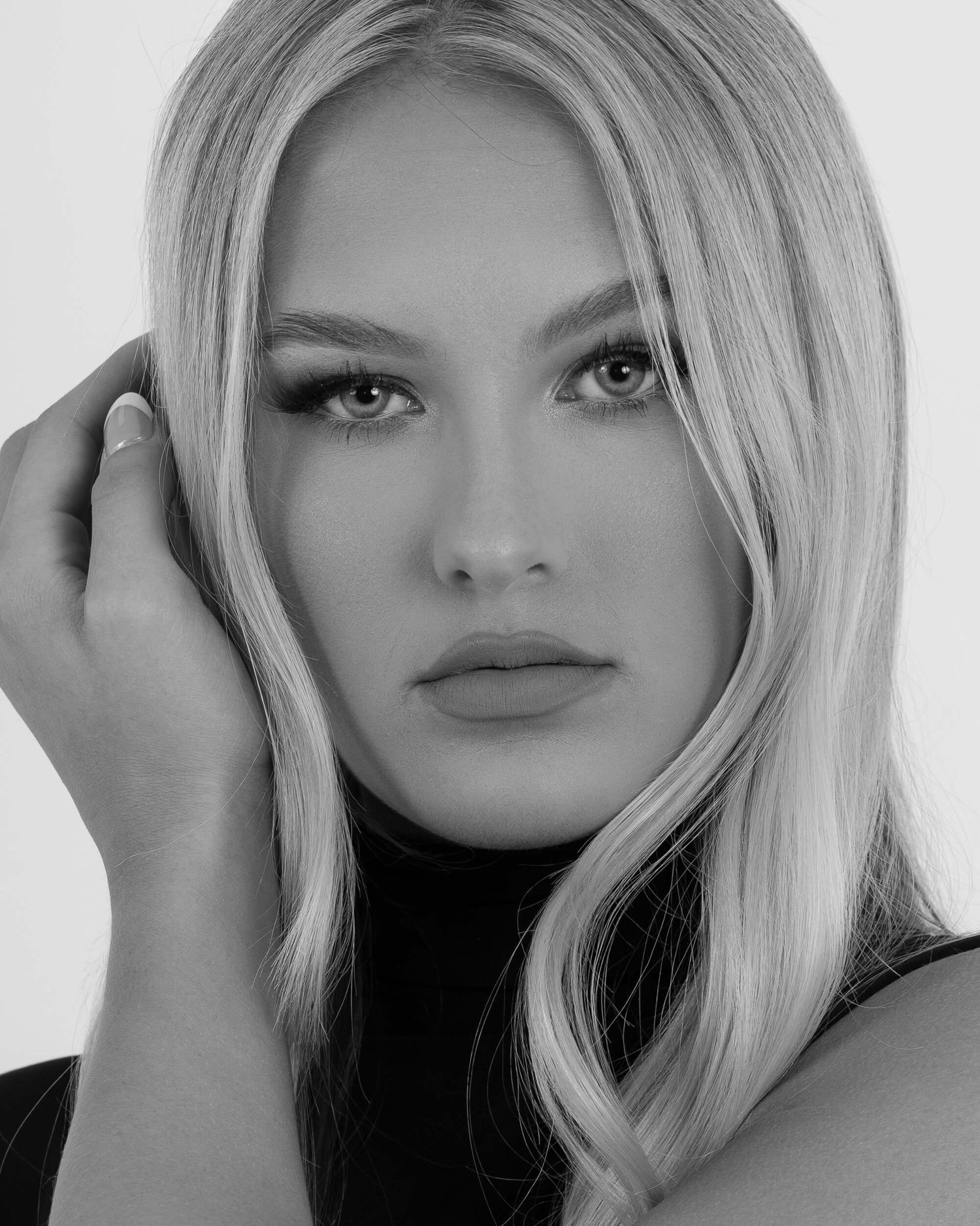 Black and white close-up portrait of a blonde woman with striking features touching her hair.