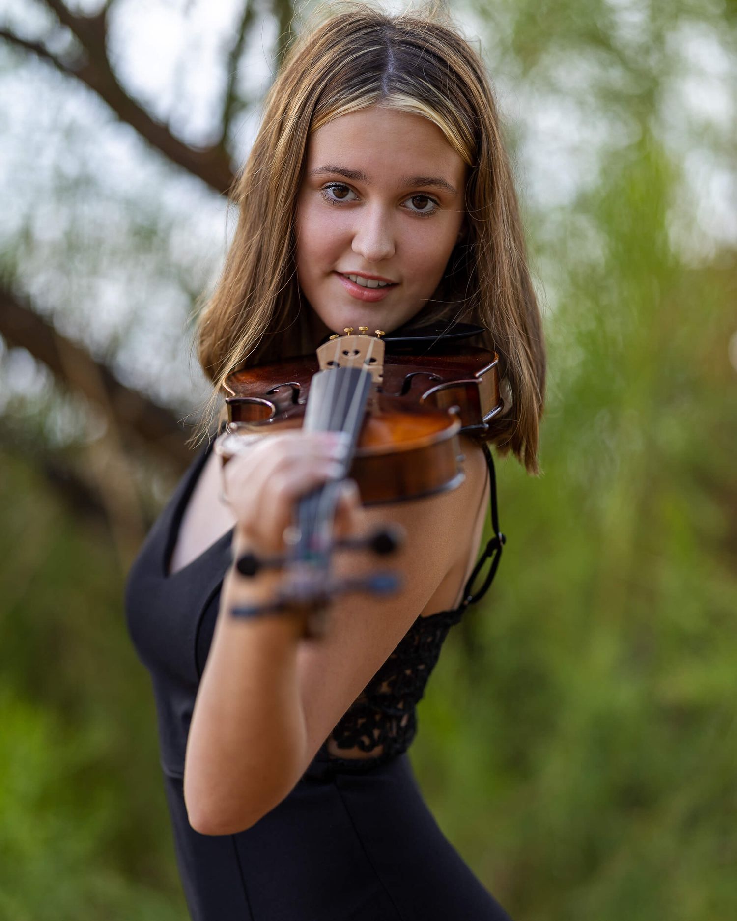 Young woman playing violin outdoors in black dress with green bokeh background.