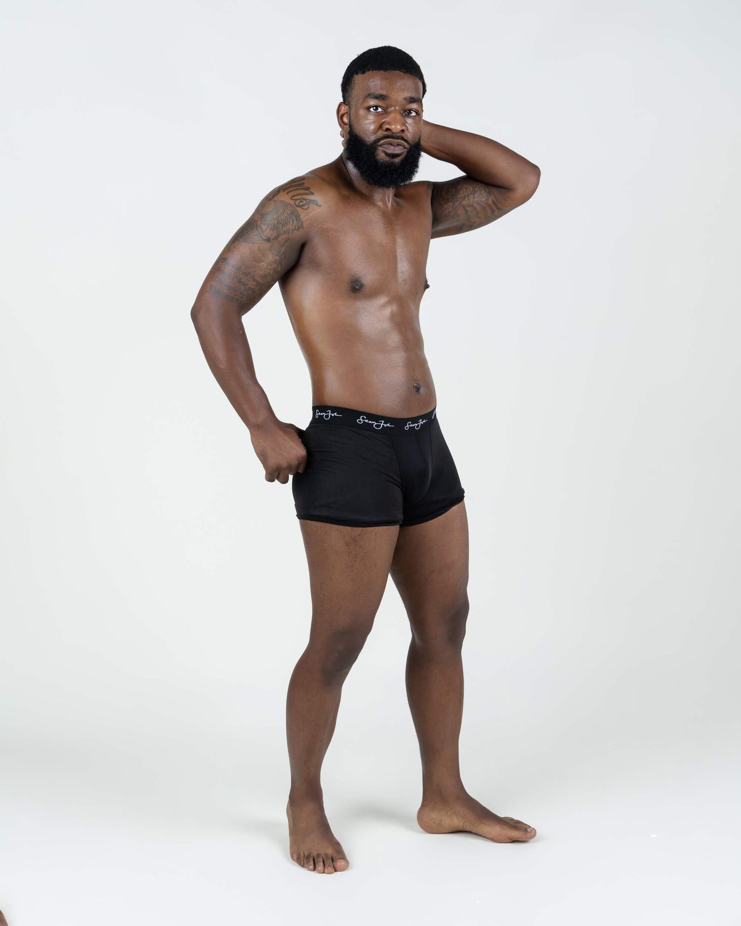 Athletic Black man with beard posing in black boxer briefs, showcasing muscular physique with tattoos.