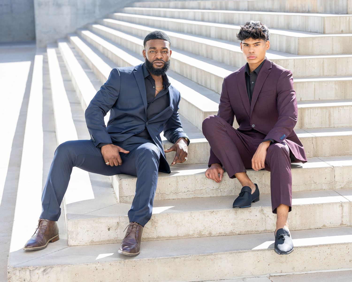 Two well-dressed men in suits sitting on outdoor concrete steps in urban setting.