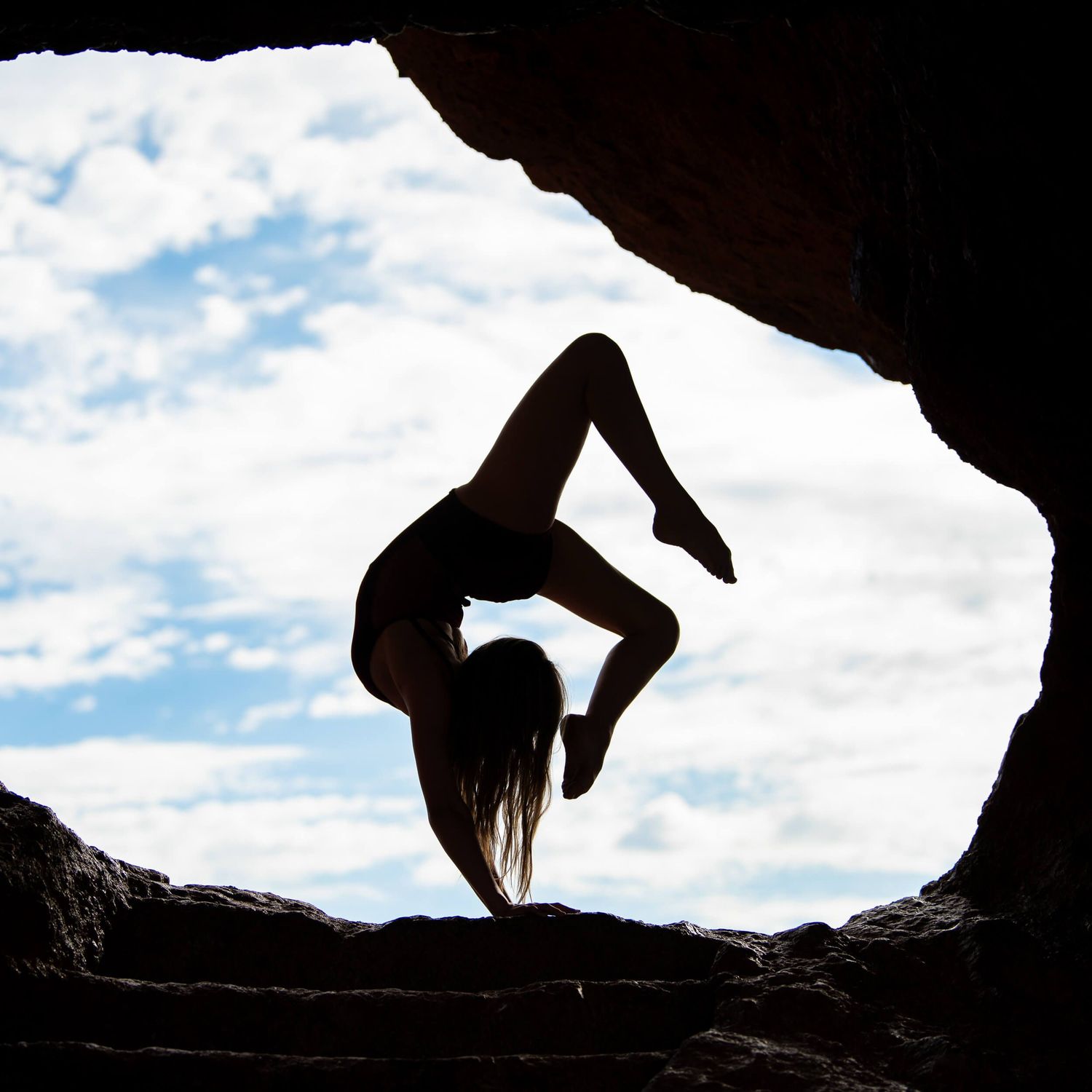 Silhouette of flexible dancer performing backbend in rocky cave opening against blue sky.