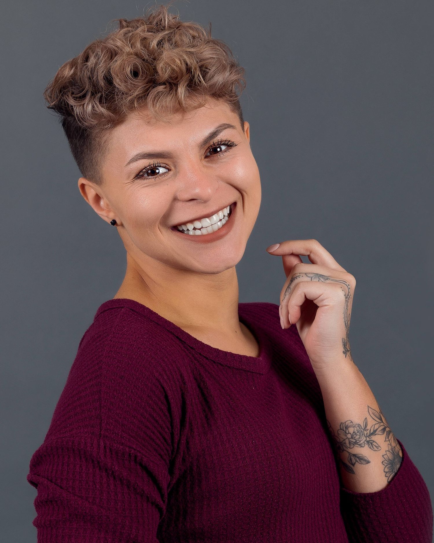 Woman with short curly hair and tattoos smiling in a burgundy top for a professional headshot.