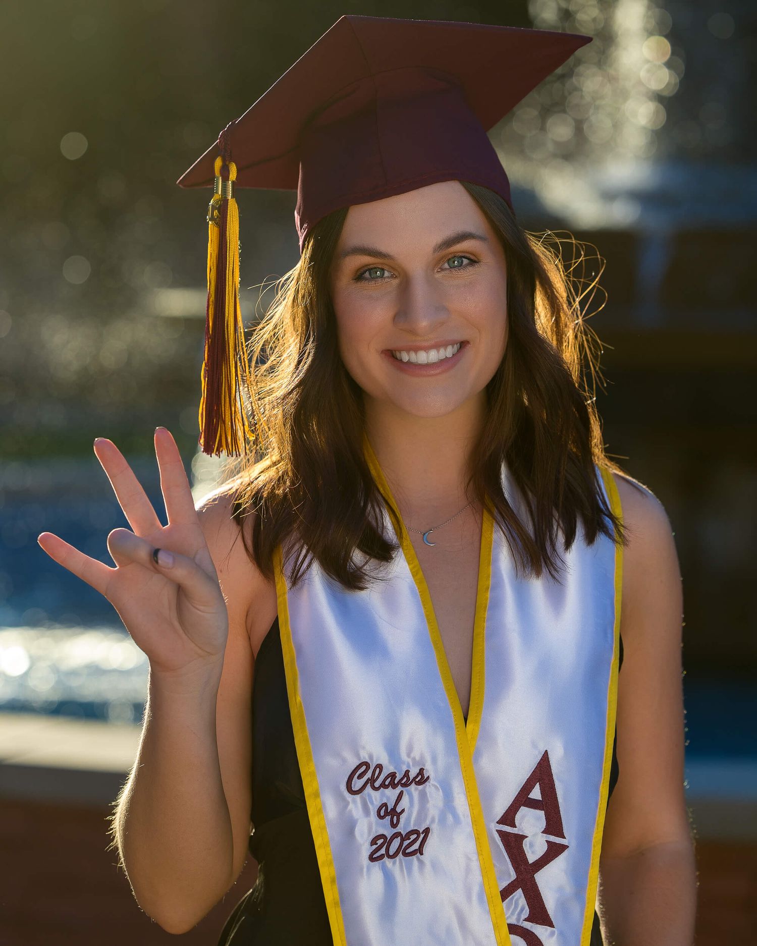 Smiling graduate in maroon cap holding peace sign wearing Alpha Chi Omega Class of 2021 stole outdoors.