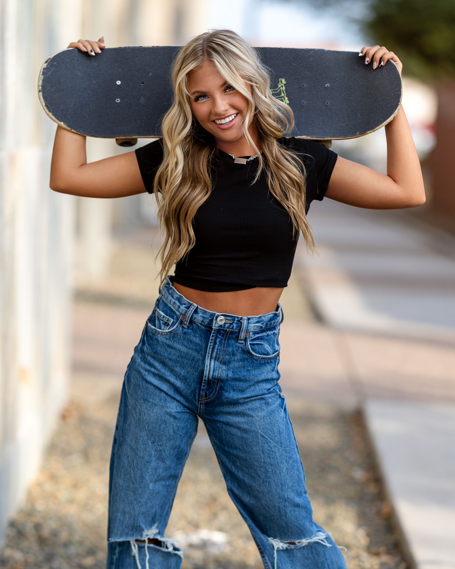 Smiling blonde woman holding skateboard behind shoulders wearing black crop top and ripped jeans.