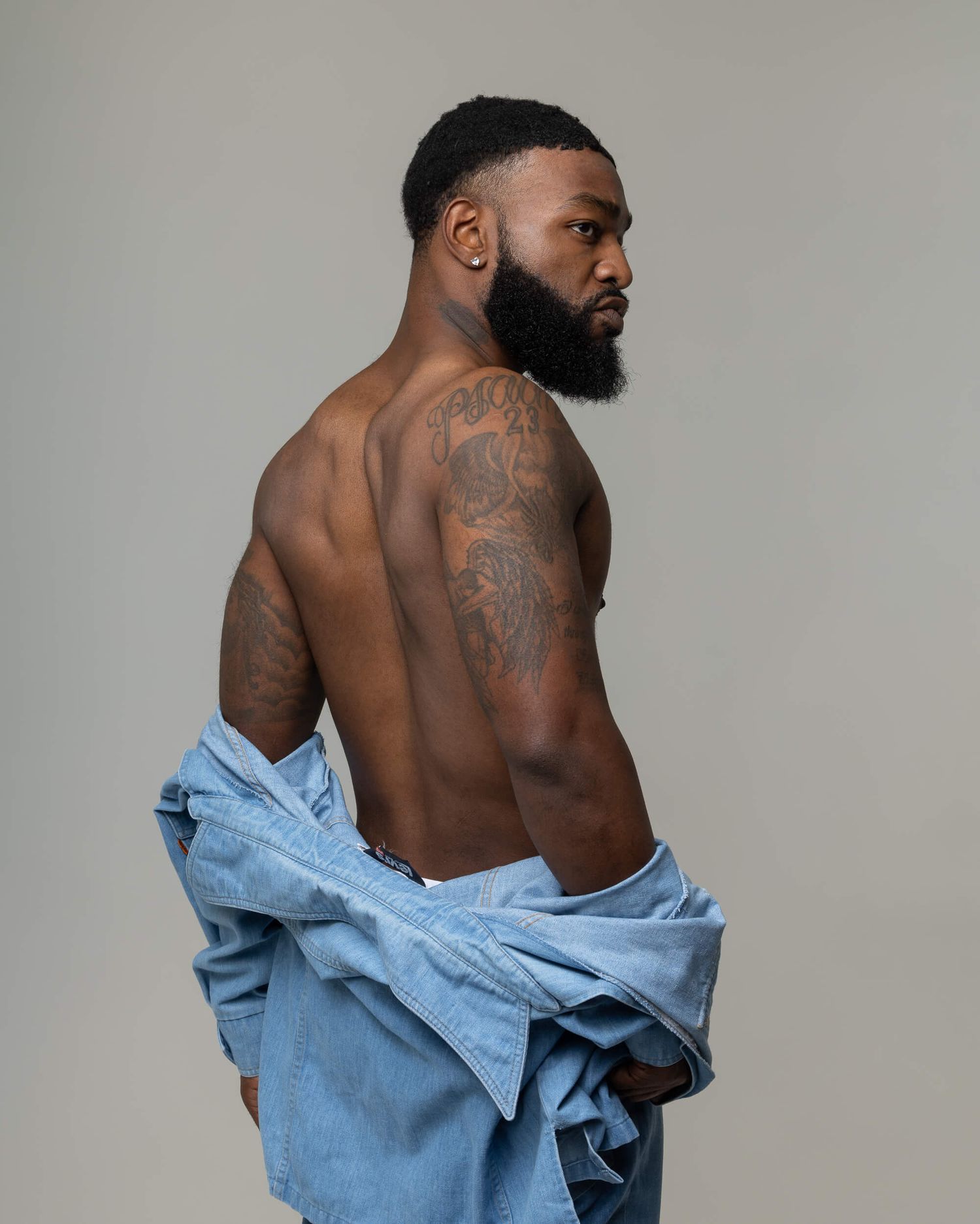 Muscular Black man with back tattoos posing shirtless with a denim jacket draped over his shoulders against a gray background.