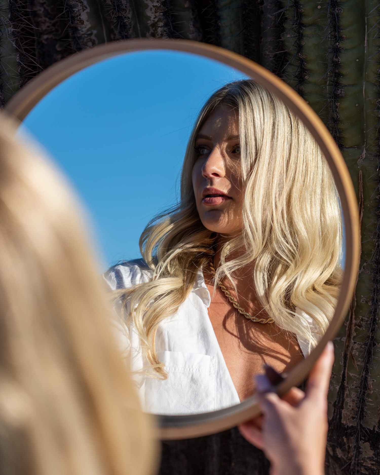 Blonde woman with curly hair reflected in round mirror outdoors.