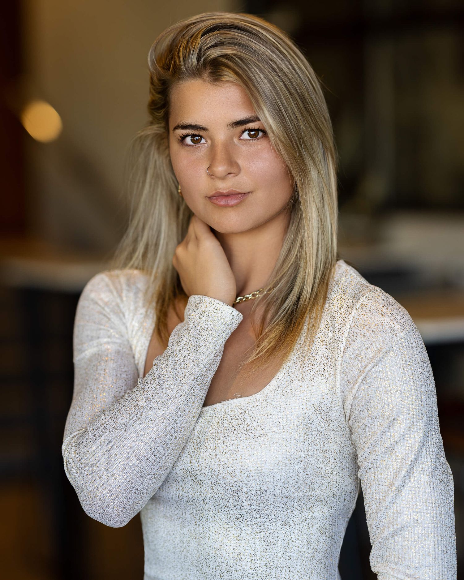 Blonde woman in white shimmer top poses thoughtfully in softly lit indoor setting.
