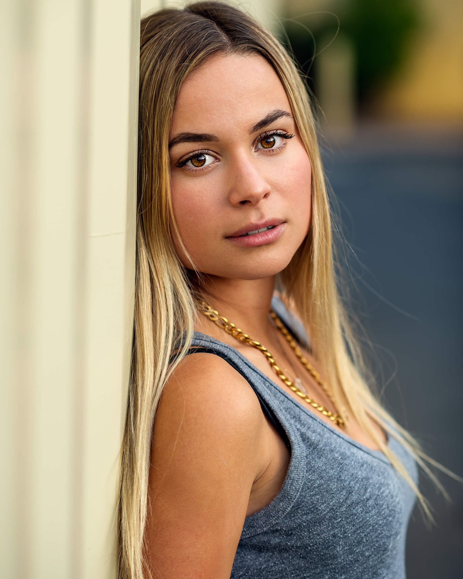 Young blonde woman with brown eyes leans against a pillar wearing a gold chain necklace.