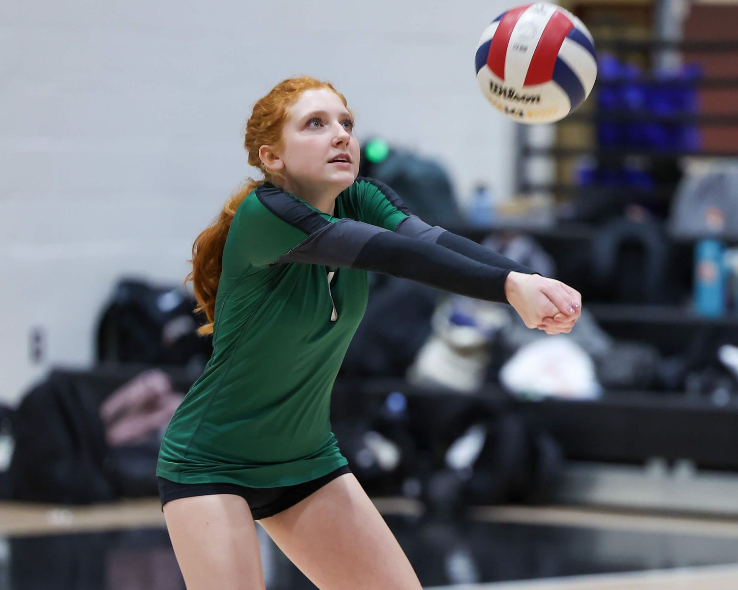 Red-haired volleyball player in green uniform bumps ball during indoor match.