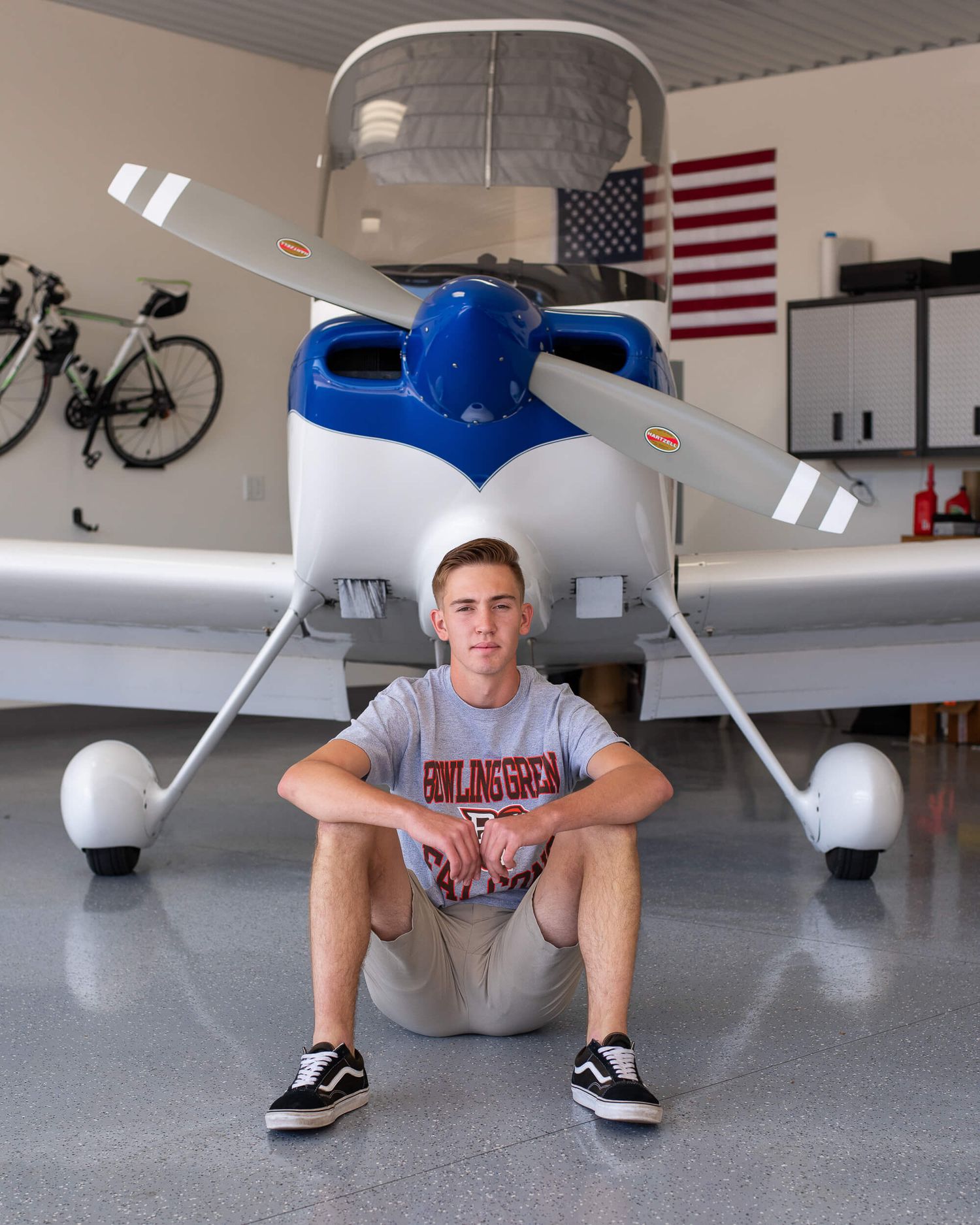 Teen boy in Bowling Green shirt sits in front of small white and blue Cessna aircraft in a private hangar.