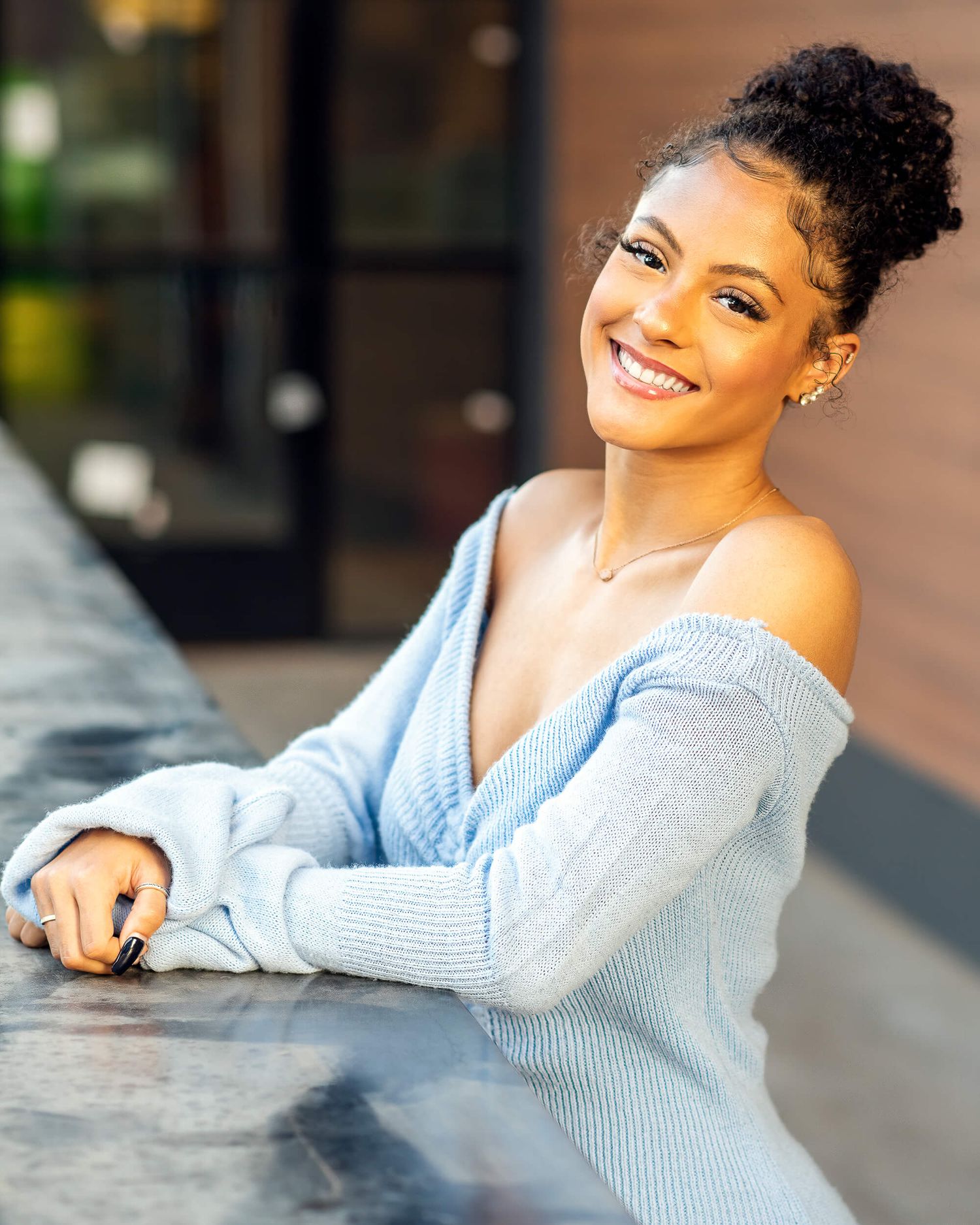 Smiling young woman with curly updo wearing off-shoulder light blue sweater, leaning on outdoor ledge.
