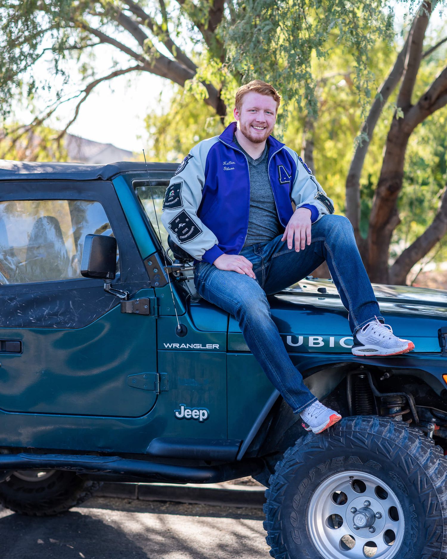 Smiling young man sitting on teal Jeep Wrangler Rubicon outdoors under trees.