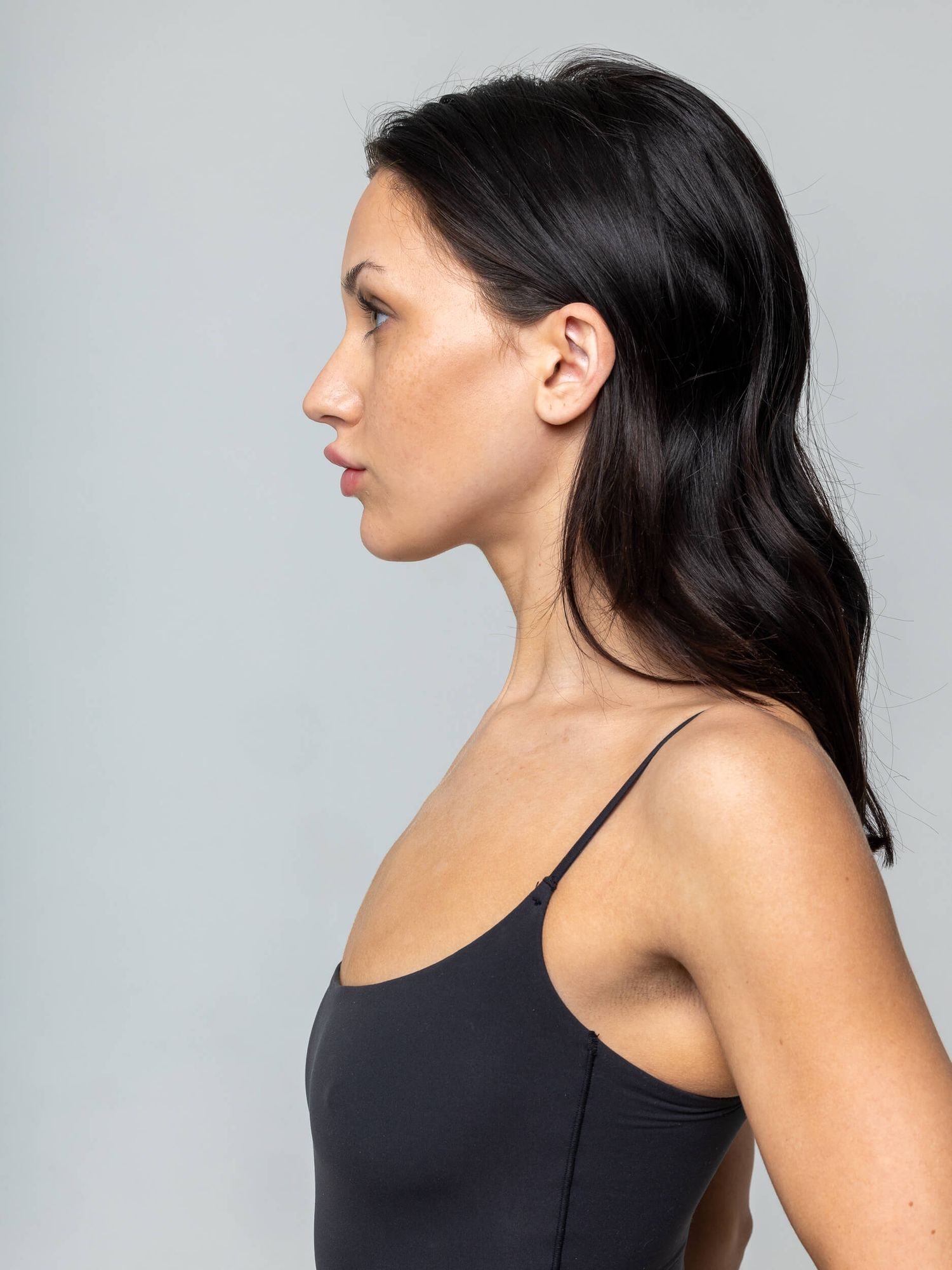 Side profile of a woman with dark hair wearing a black spaghetti strap bodysuit against a gray background.