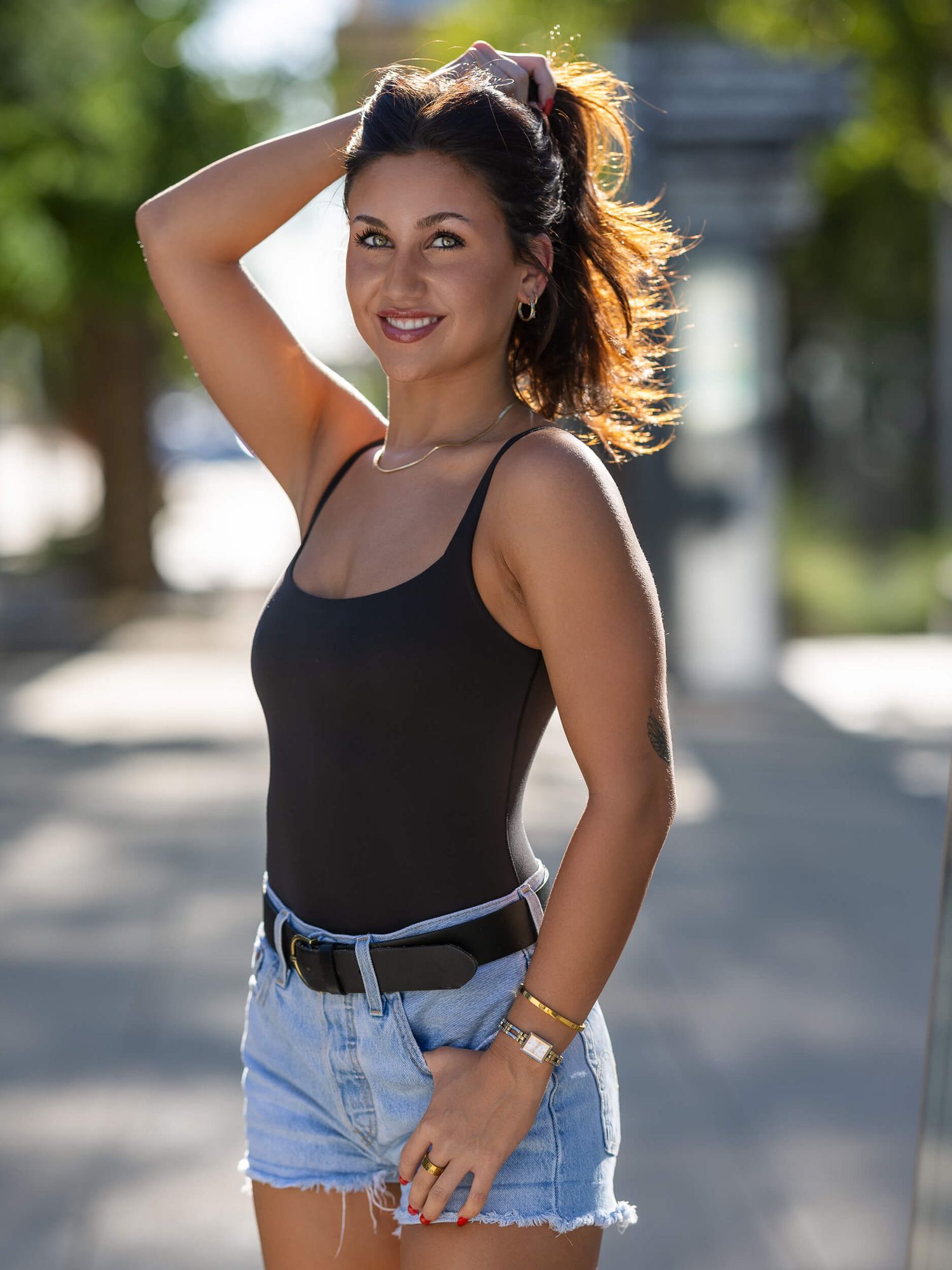 Smiling woman in black tank top and denim shorts posing outdoors on a sunny day with hair blowing in the wind.