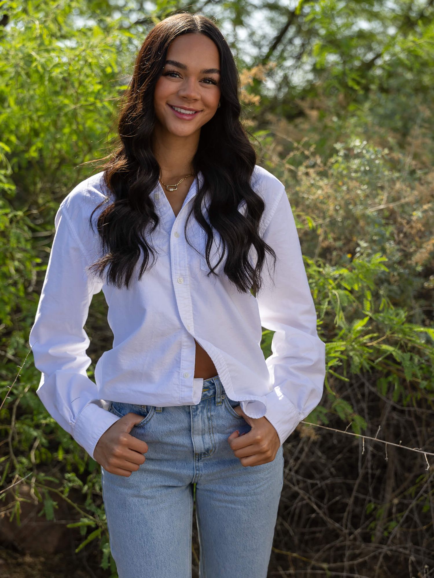 Young woman smiling in white cropped button-up shirt and light jeans outdoors.