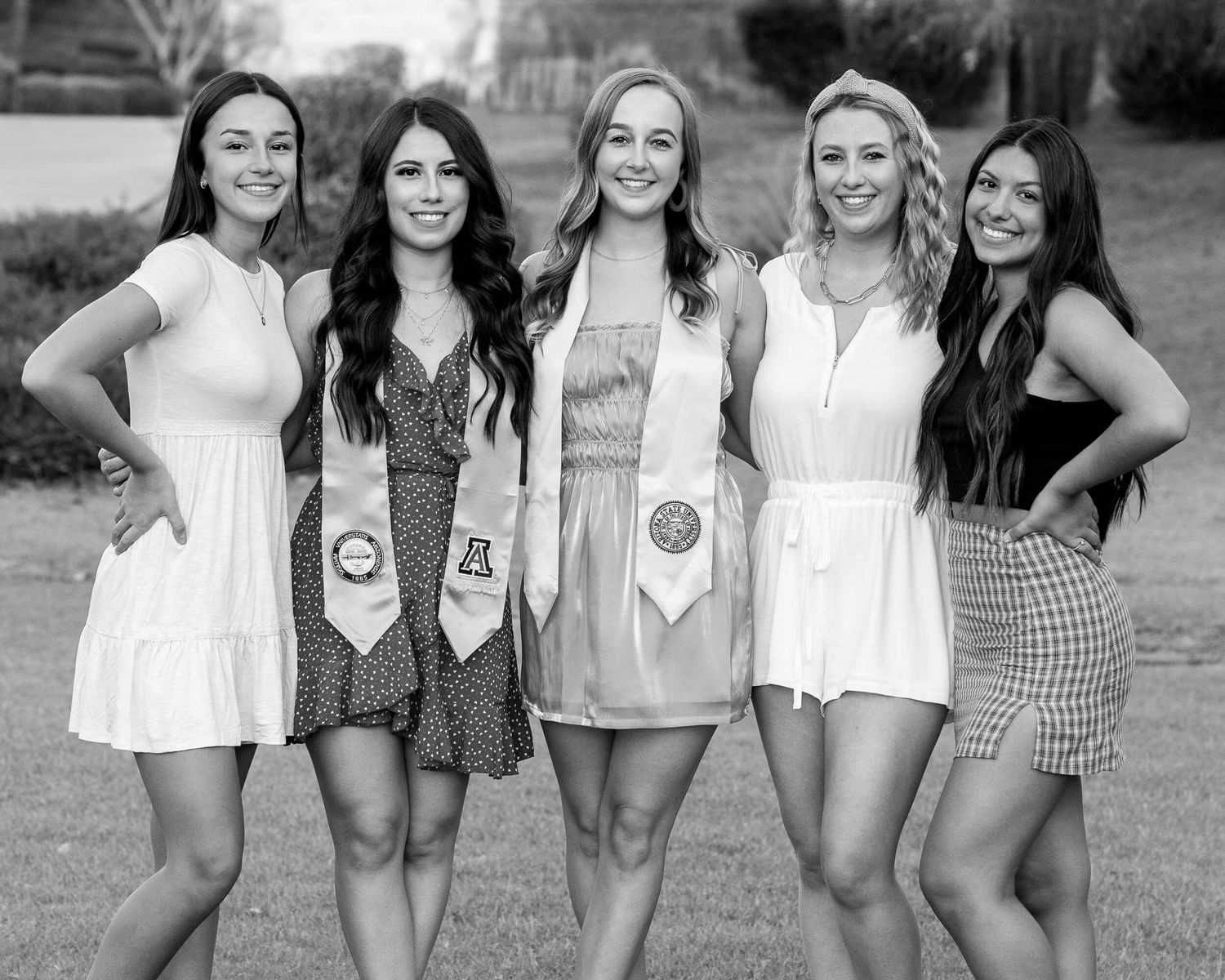Black and white photo of five smiling young women in graduation stoles posing outdoors.
