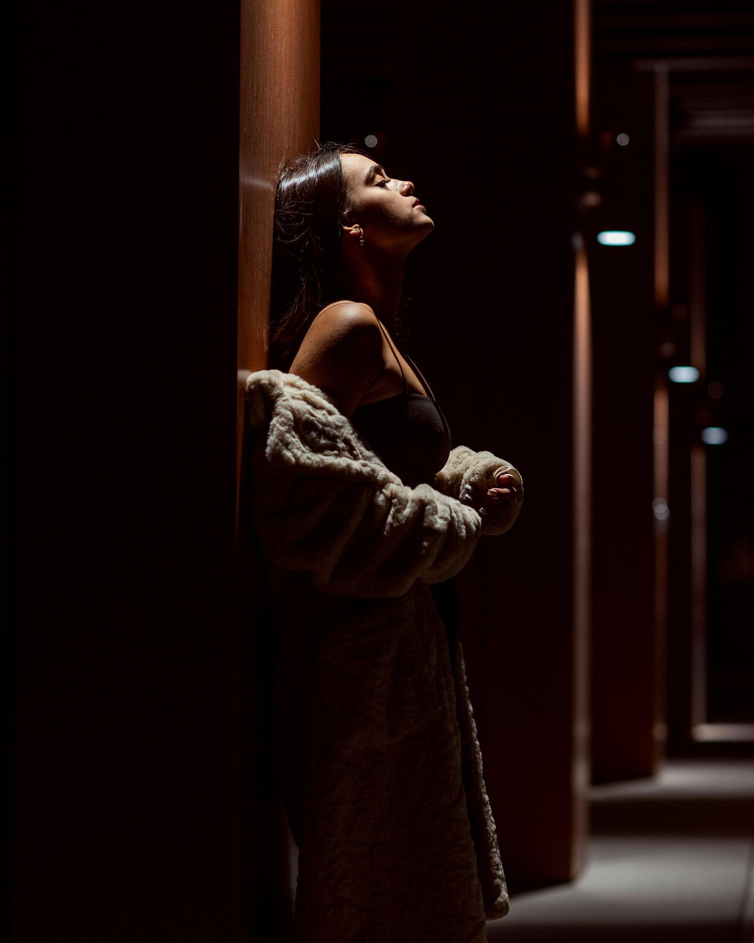 Woman in fur coat leaning against wooden pillar in dark hallway.