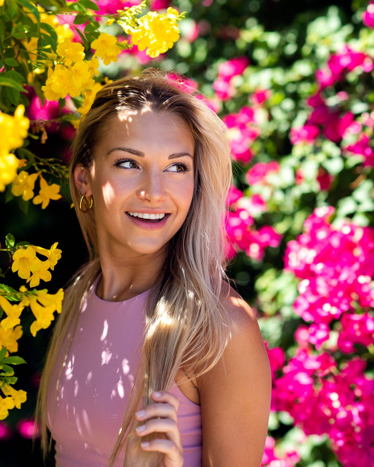 Smiling blonde woman in pink top surrounded by vibrant yellow and pink flowering bushes.