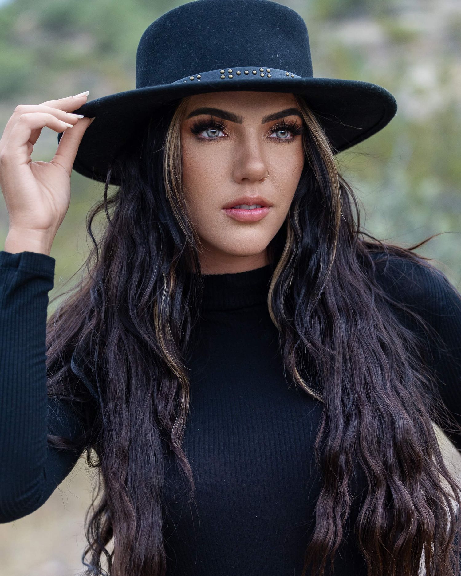 Striking brunette woman with green eyes wears a black wide-brim hat and black top.