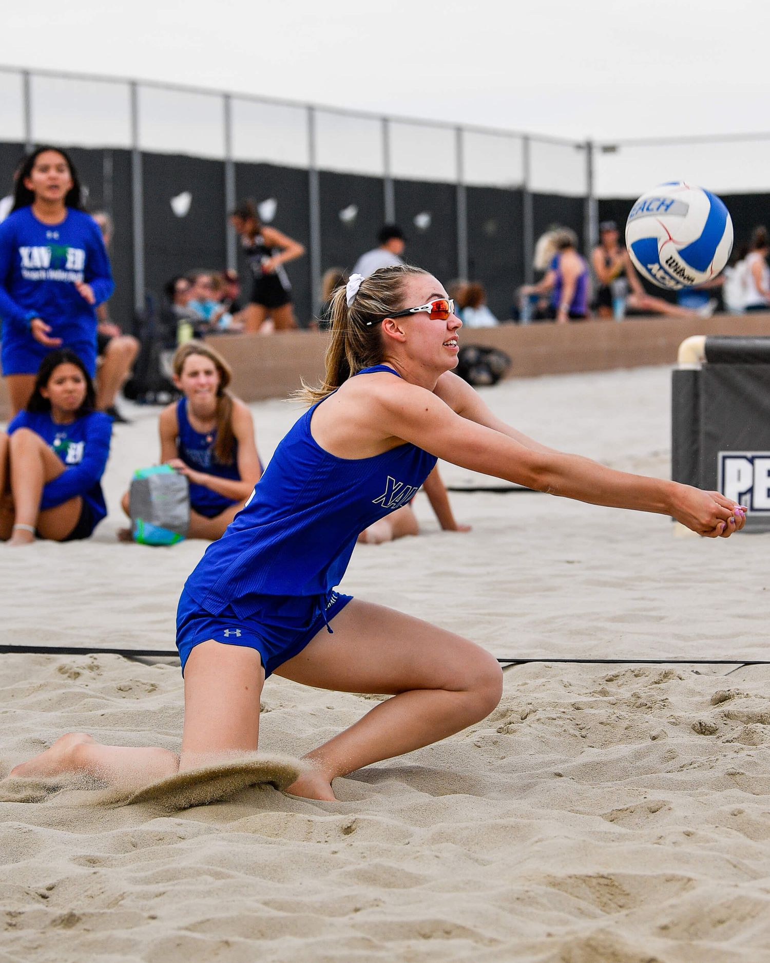 Female volleyball player in blue uniform diving to dig a ball on a sandy beach volleyball court.