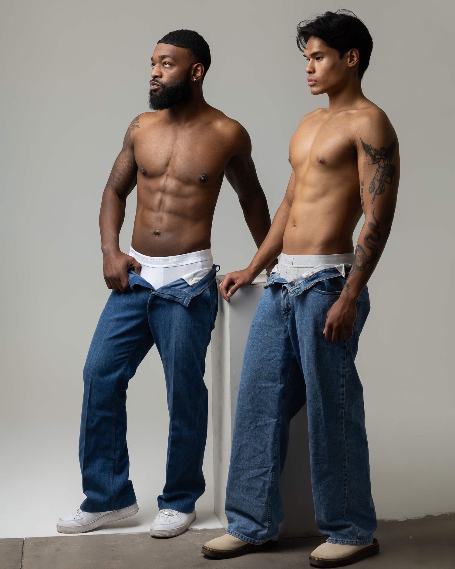 Two shirtless athletic men in low-rise blue jeans posing together in a studio with white background.