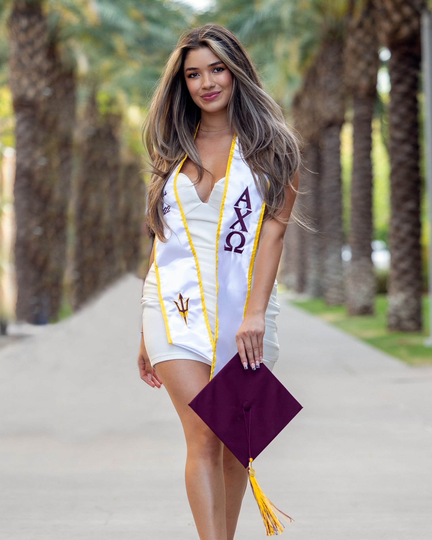 Woman in white dress wearing Alpha Chi Omega graduation stole holding maroon cap near palm trees.