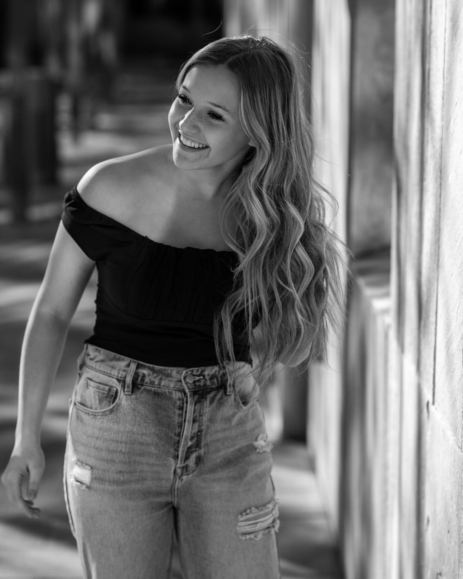 Smiling blonde woman in black off-shoulder top and jeans, black and white photo.