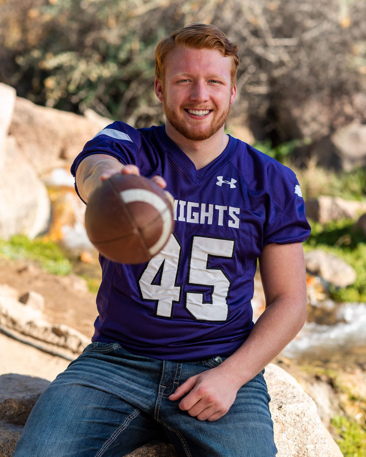 A smiling young man in a purple Knights football jersey number 45 holds a football toward the camera outdoors.