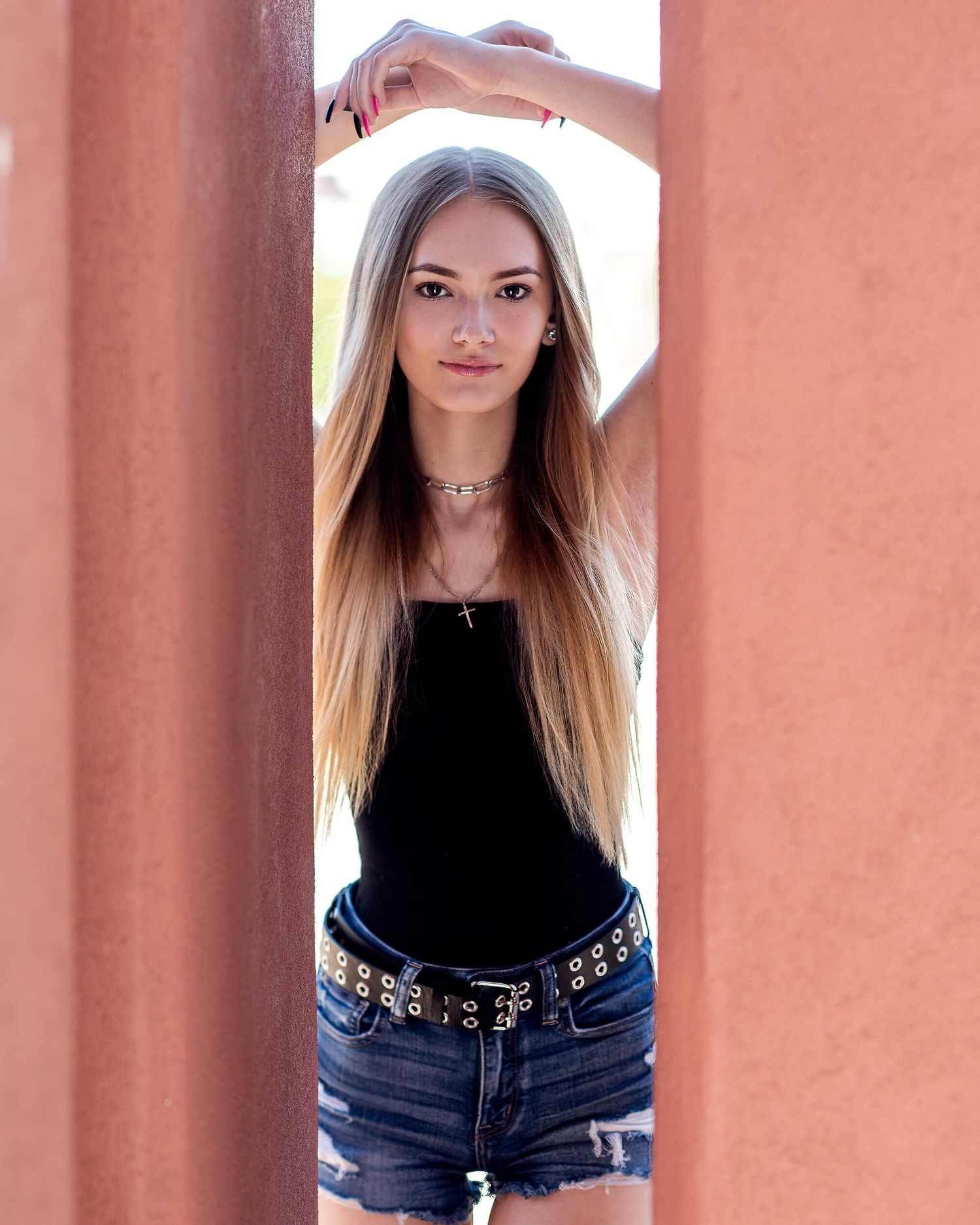 Young blonde woman in black tank top and denim shorts posing between two rust-colored pillars outdoors.