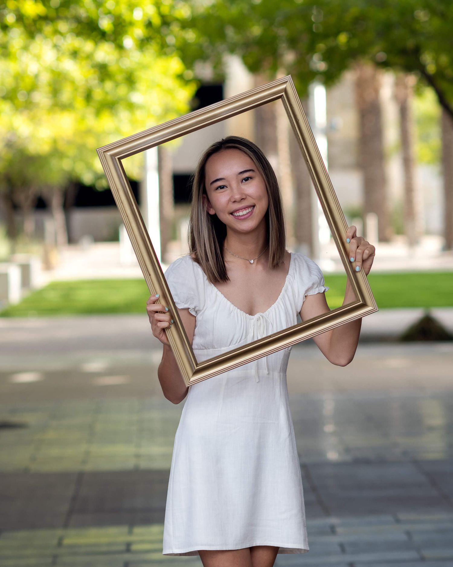 Smiling young woman in white dress holds golden picture frame outdoors in park setting.