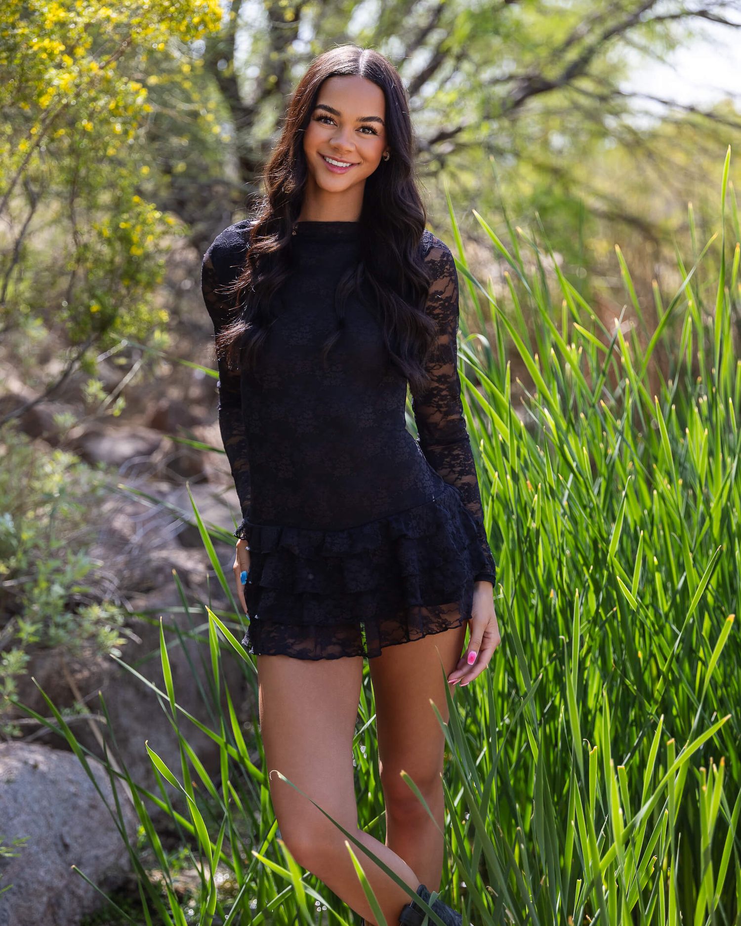 Woman in black lace mini dress smiling outdoors among tall green grass and plants.