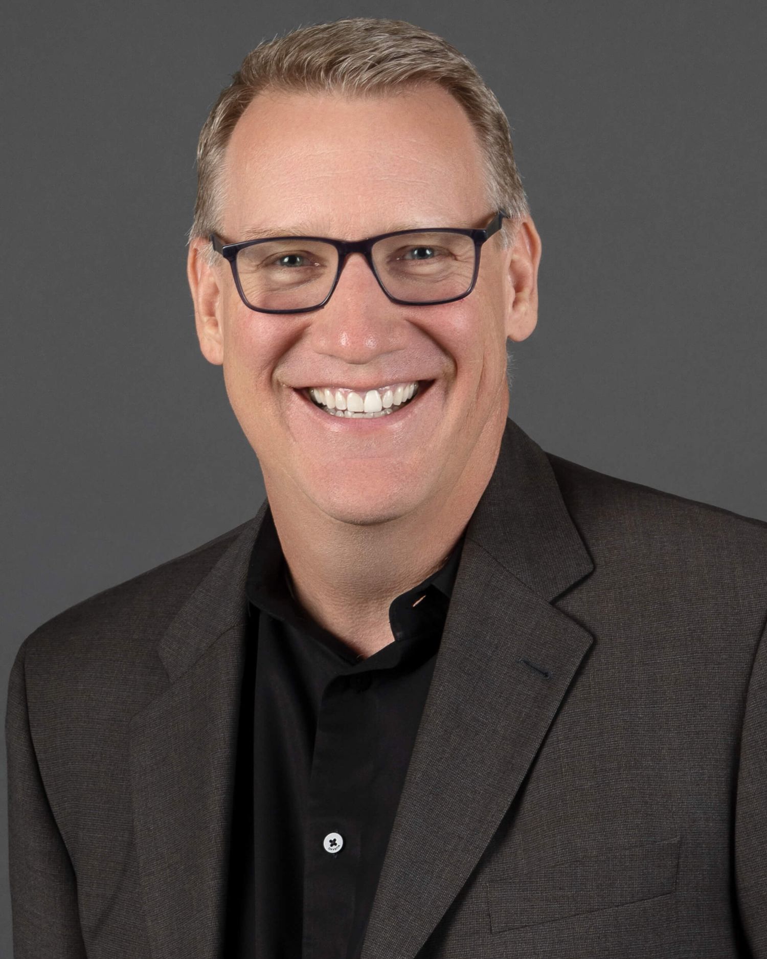 Professional headshot of a smiling middle-aged man with gray hair and glasses wearing a black shirt and gray blazer.
