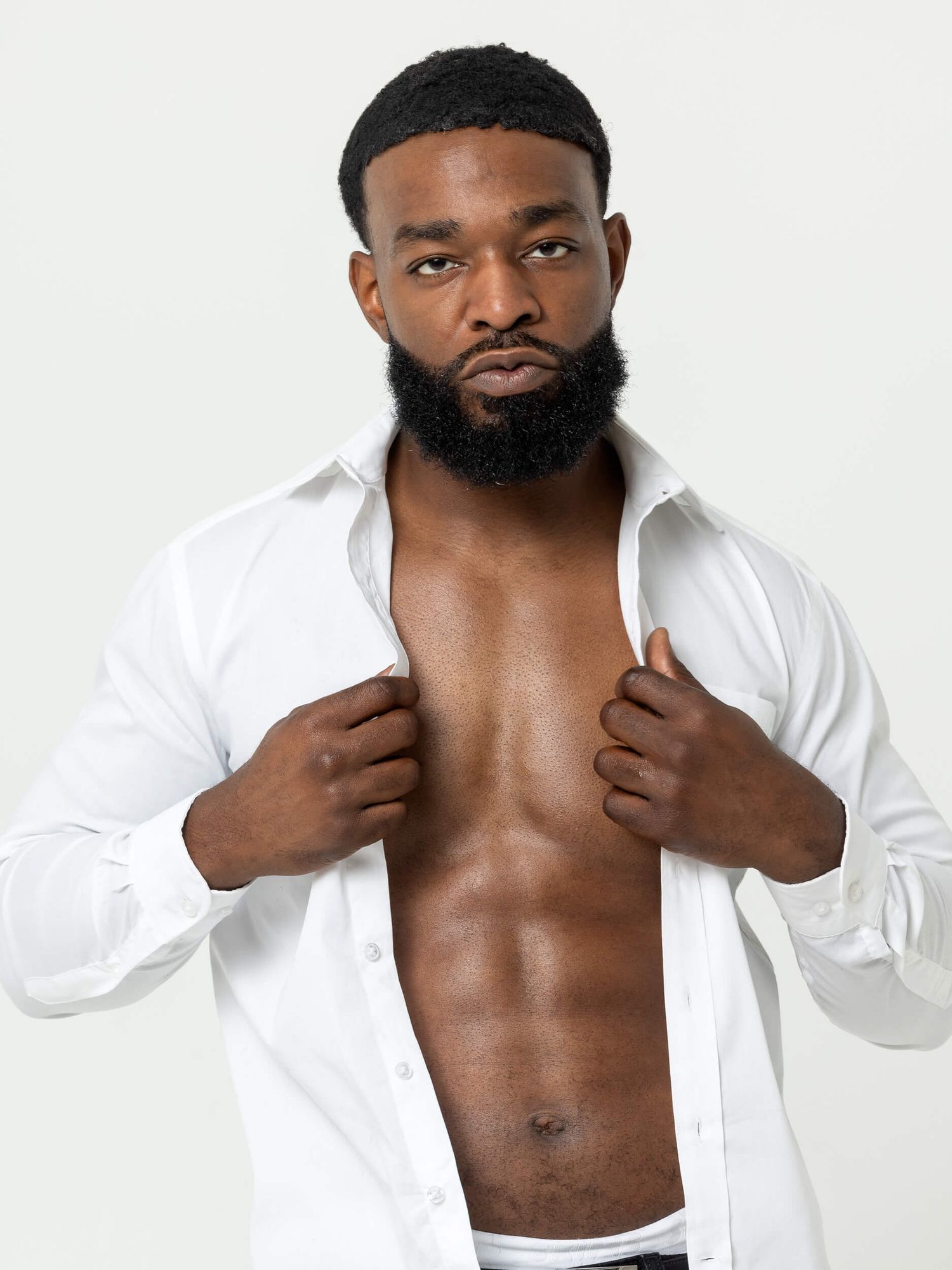 Muscular Black man with beard posing in open white dress shirt showing toned abs against light gray background.