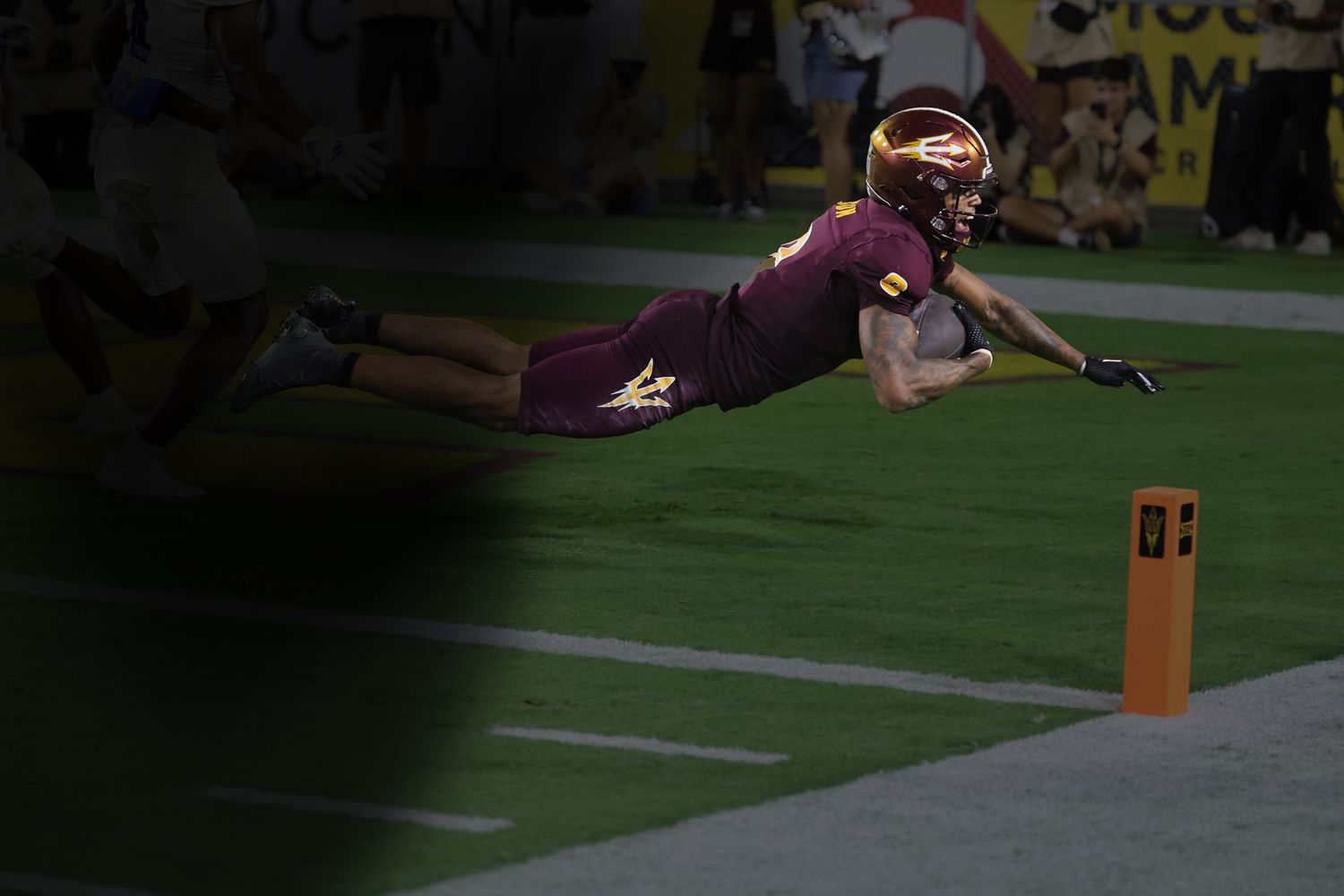 Football player dives horizontally toward the end zone pylon during a night game.