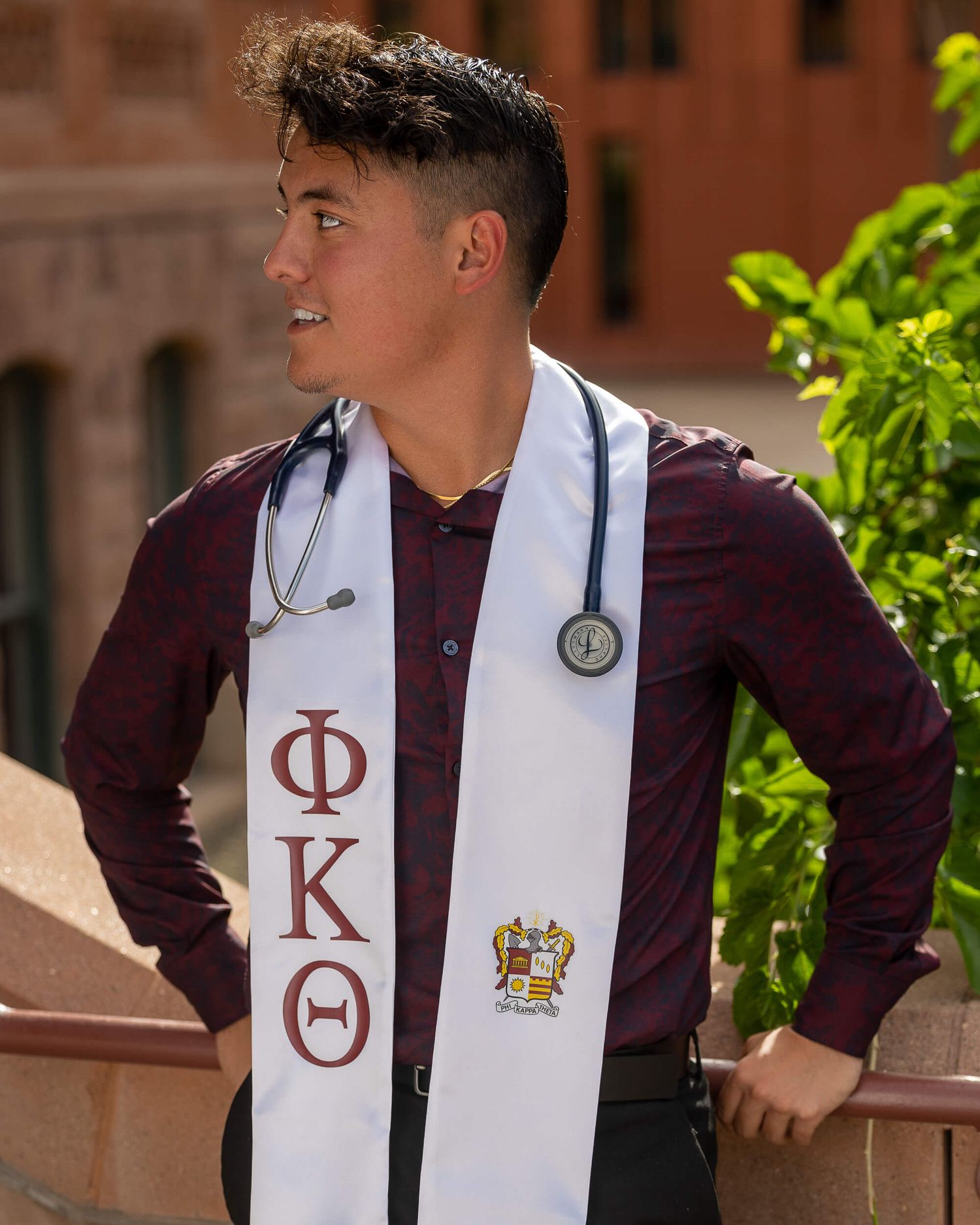 Male graduate wearing Phi Kappa Theta stole and stethoscope poses at a brick campus building.