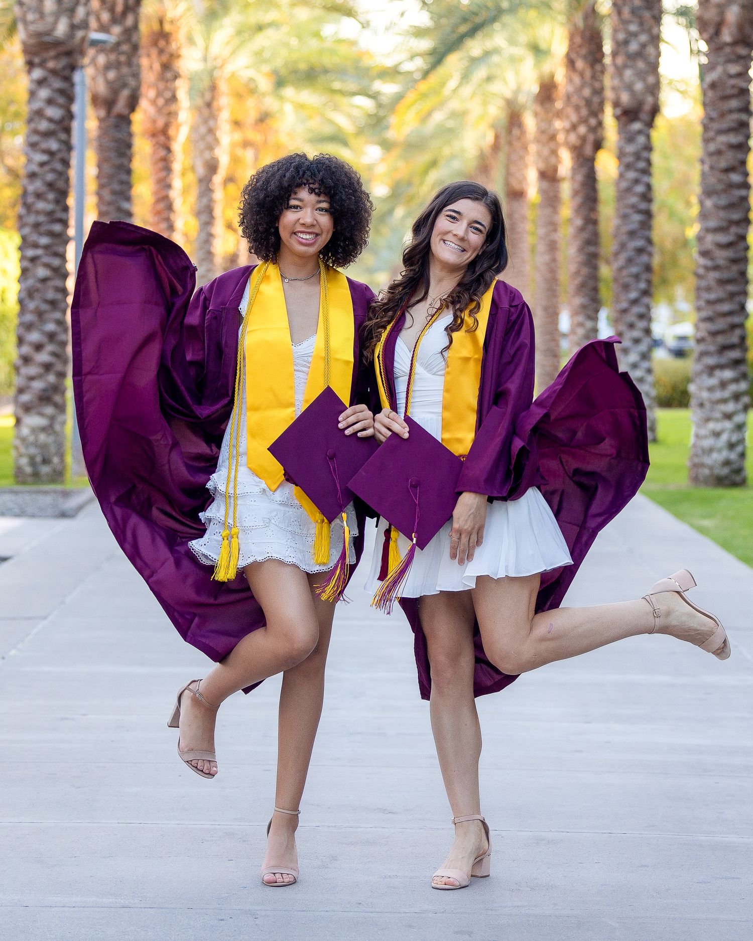 Two graduates in maroon caps and gowns jump joyfully among palm trees on campus.