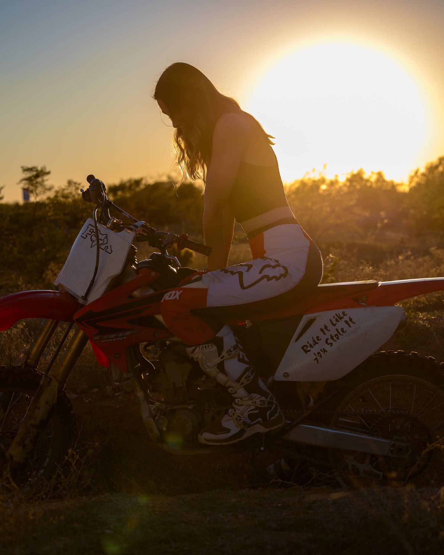 A female motocross rider sits on a red dirt bike during a stunning golden sunset outdoors.