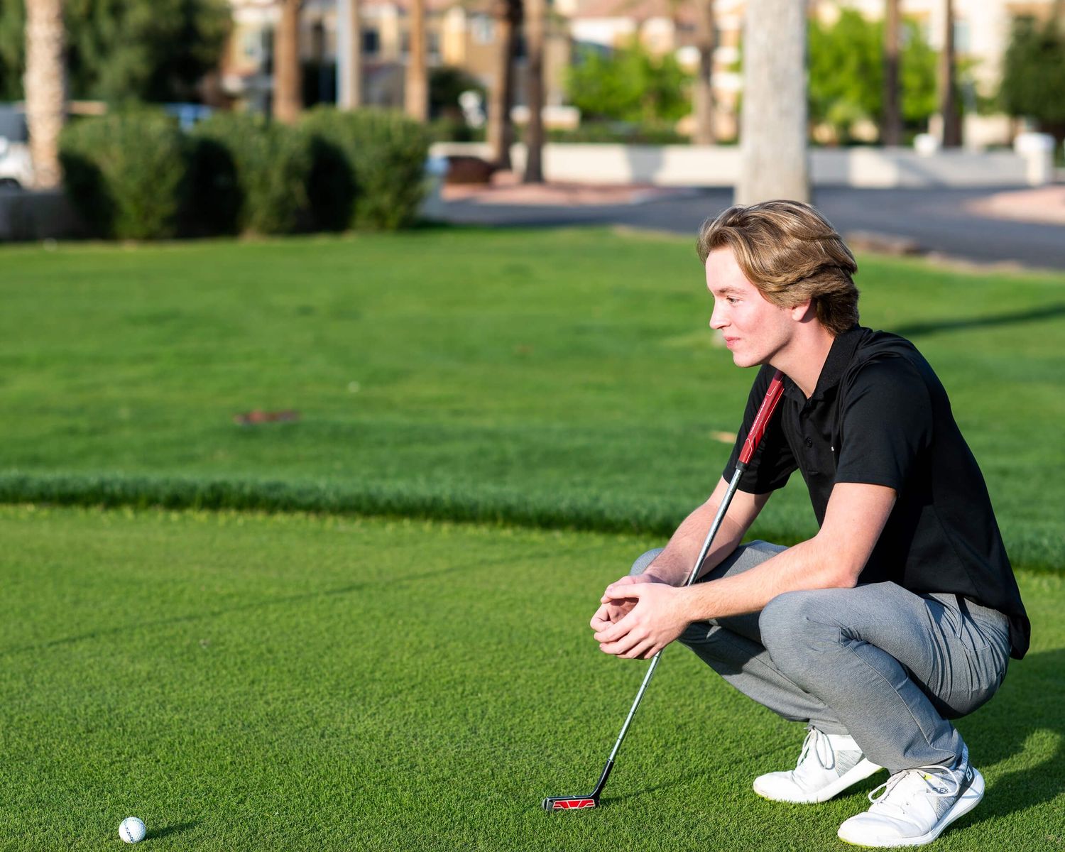 Young golfer crouches to read green while holding putter at sunny golf course.