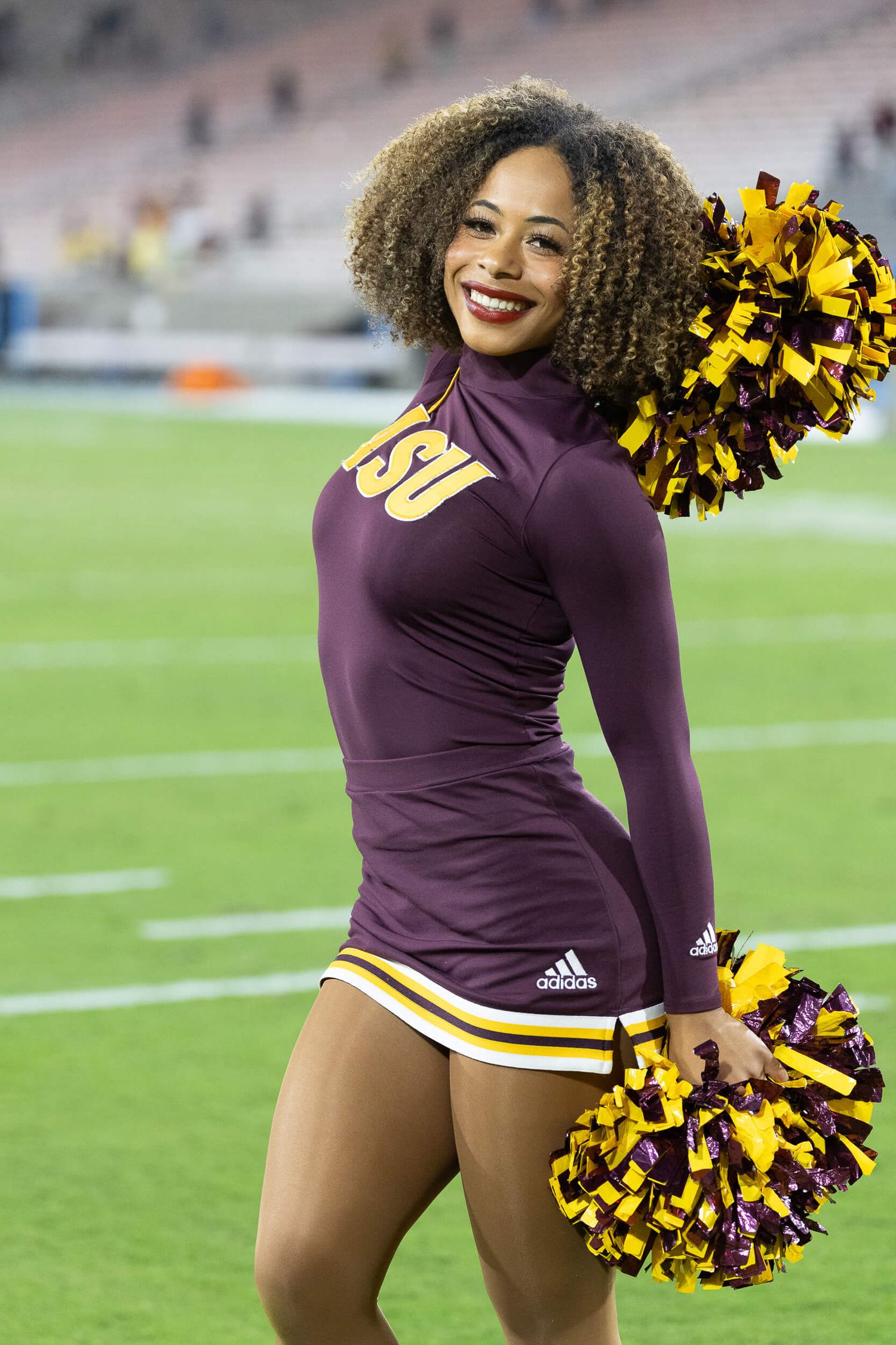 ASU cheerleader with curly hair smiles in maroon Adidas uniform holding gold and maroon pom-poms on the field.
