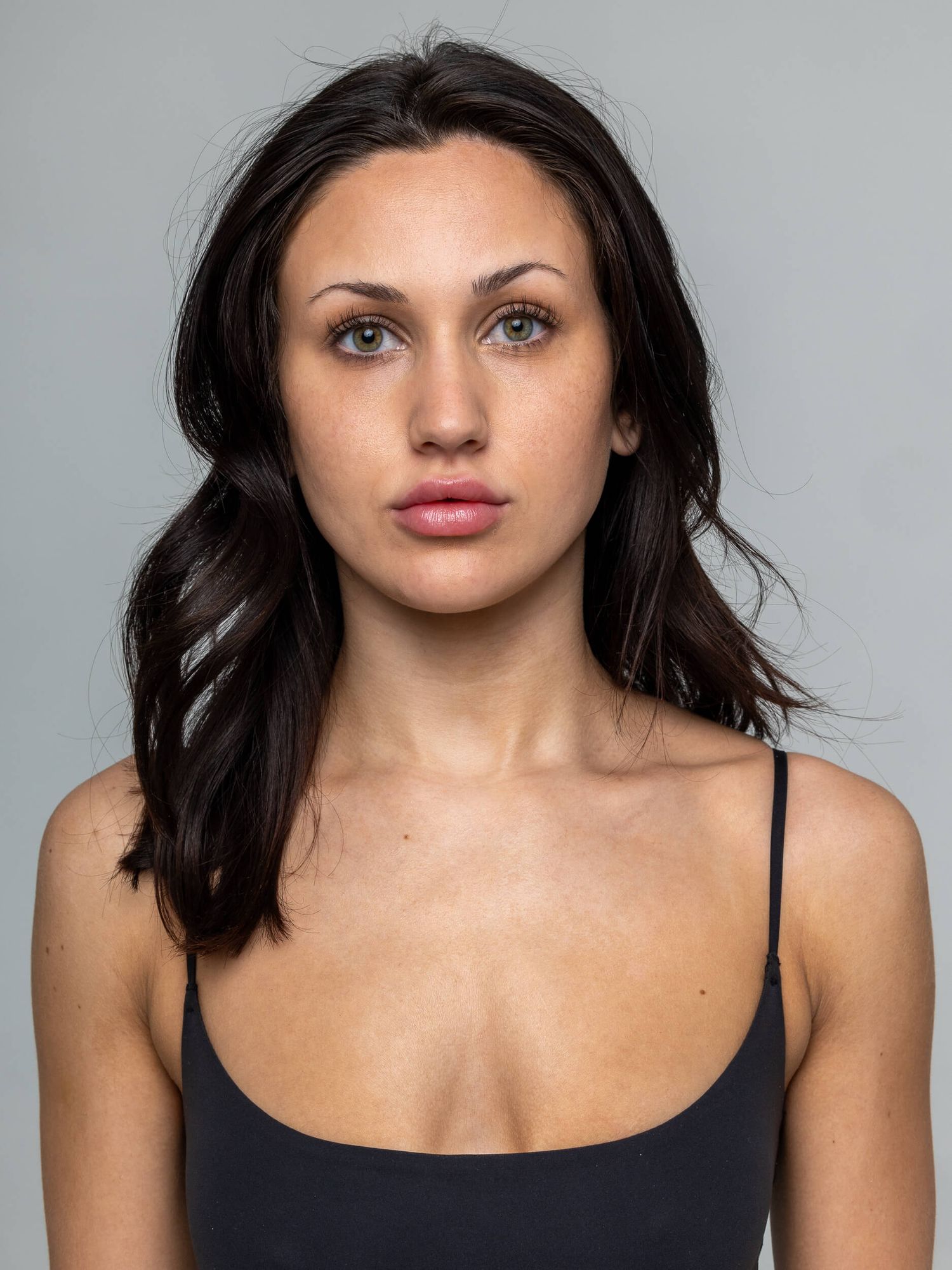 Young woman with dark hair and green eyes wearing a black tank top, posing against a gray background.